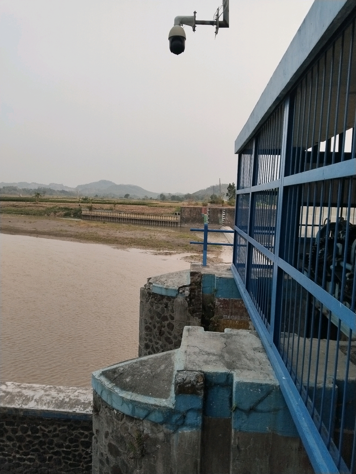 The image depicts a view of a dam or water control structure in Prupuk Utara, Indonesia. In the foreground, a concrete and stone structure with blue metal railings is visible. This structure appears to be part of a walkway or maintenance area overlooking a body of murky brown water. A security camera is mounted on a metal pole above this structure, pointing downwards. The background shows a landscape with agricultural fields and rolling hills under an overcast sky. There are no people visible in the image.