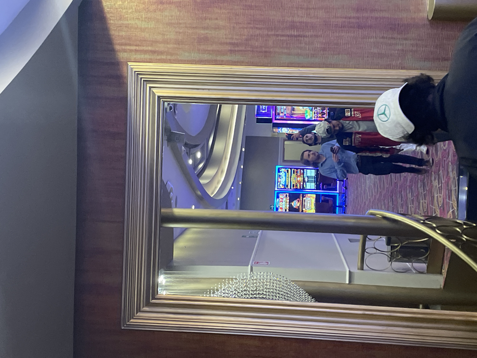 The image shows a framed mirror in a room with patterned wallpaper and a grey wall to the side. The mirror reflects a casino interior, including slot machines, a curved ceiling design with lighting, and a patterned carpet. Two people are reflected in the mirror standing in the casino. One is wearing a white baseball cap with a Mercedes logo and the other is wearing a black and red shirt and white baseball cap with writing. The mirror also shows part of a golden-colored decorative fixture. It is likely that this image was taken inside a casino in Huixquilucan de Degollado, Mexico.
