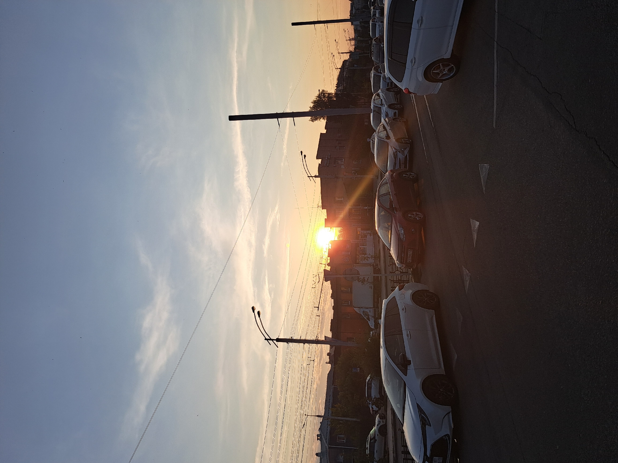 The image captures a sunset scene on a city street, likely in Irkutsk, Russia, based on the context provided. The sky is a gradient of blue and orange, with the sun visible on the right side of the frame, casting a warm glow. A few thin clouds are present in the sky. Several vehicles are lined up on the road, indicating traffic. On the left, there are buildings and trees. Overhead, power lines crisscross the sky, supported by utility poles. One prominent pole with streetlights stands near the center of the image. The overall impression is of a busy urban environment at dusk, with the natural beauty of the sunset juxtaposed against the man-made structures and activity. There are no visible people in the image.