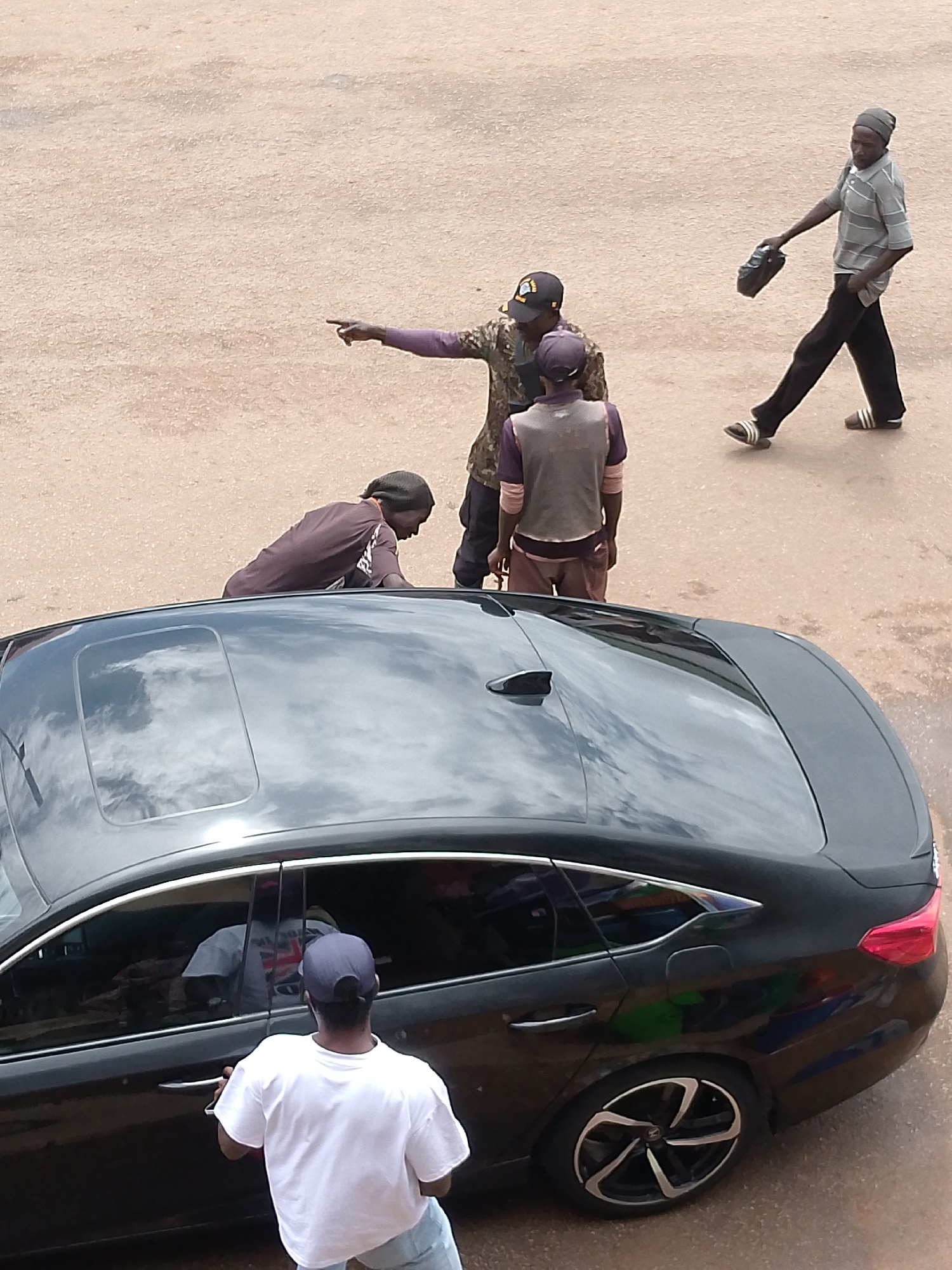 In Bukuru, Nigeria, an overhead view captures a street scene with several individuals and a black sedan. One person, wearing a grey and white striped t-shirt, dark pants, and a grey beanie, walks towards the left, holding a black bag. Nearby, three other individuals are gathered. One, with a camouflage patterned shirt and a dark baseball cap, points towards the left with his right arm. Another person, in a purple long-sleeved shirt and a grey vest, stands facing the car. A fourth person, wearing a dark purple t-shirt and a dark beanie, is leaning over the roof of the black sedan, possibly inspecting or interacting with the vehicle. Inside the car, a person in a white t-shirt is visible through the tinted windows. The ground is a light brown, granular surface, typical of a dirt road or a paved area with sand. The car itself is a dark, glossy sedan with a sunroof. The overall impression is a candid snapshot of everyday activity on a Nigerian street.