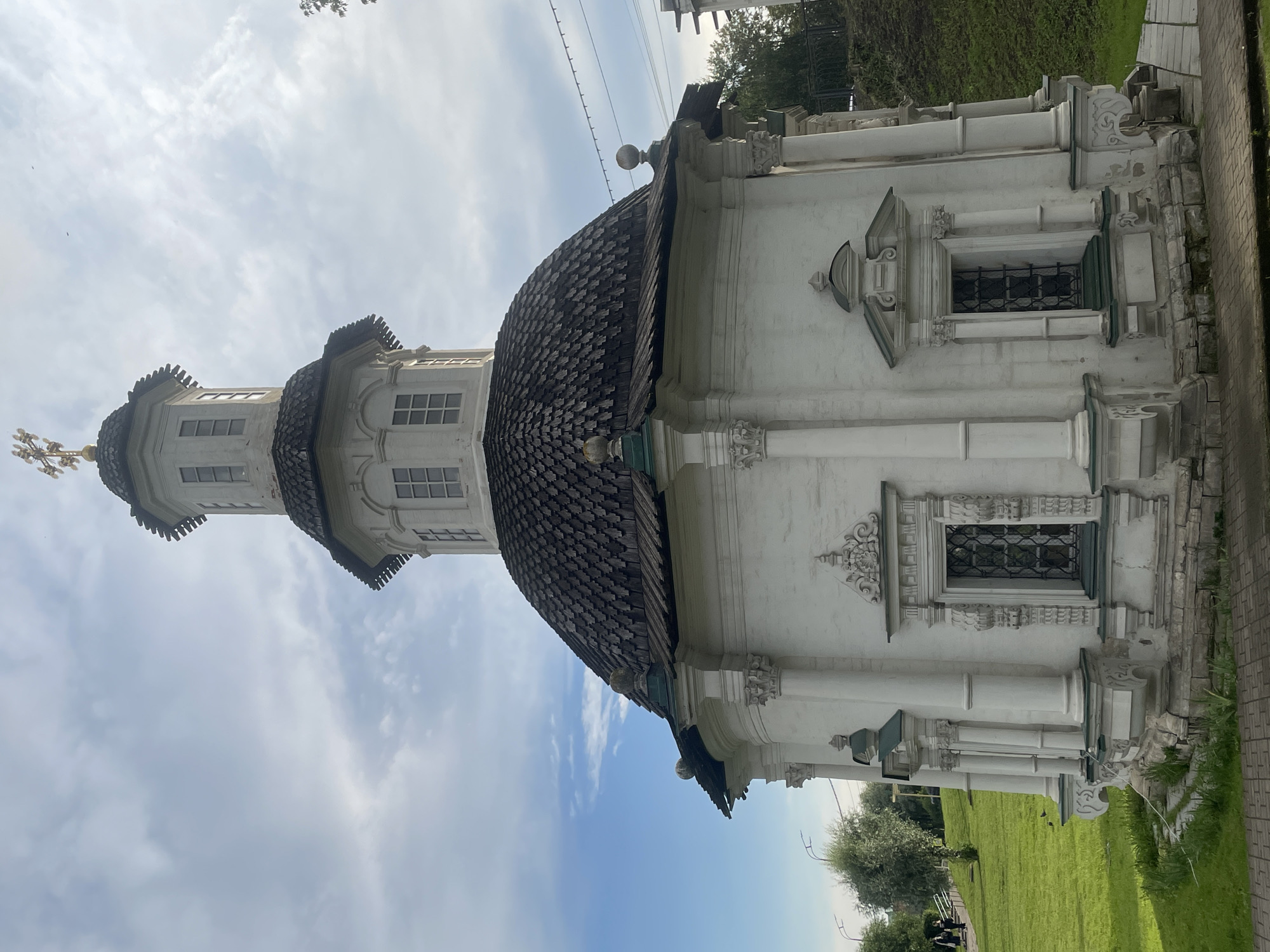 This image, taken in Sergiyev Posad, Russia, showcases a section of a white, ornate building, likely a church, with a dark, multi-layered dome. The building features classical architectural elements such as columns and decorative carvings around its windows. The windows themselves are rectangular and appear to have protective grilles. The dome is covered in dark shingles arranged in a scale-like pattern, and it is topped by a golden cross. The building's white facade has some areas where the paint appears weathered or chipped, suggesting age.
The scene is set outdoors under a bright, cloudy sky. Sunlight illuminates the building, casting subtle shadows. To the right of the building, a paved walkway runs alongside a well-maintained green lawn. In the background, trees and foliage are visible, and further in the distance, what appear to be electrical wires stretch across the sky, with some foliage from overhead trees casting shadows. At the bottom left of the image, a few individuals are visible walking on a path near the lawn and trees, appearing as small figures in the overall scene. The overall impression is of a peaceful, daytime scene in a historical or religious setting. There is no visible text in the image.