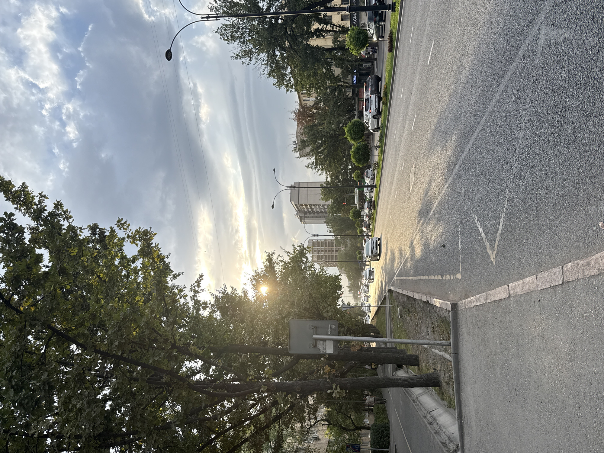 The image depicts a wide, paved road in Bishkek, Kyrgyzstan, lined with mature trees on the left side. The sun is low in the sky, casting a warm glow and creating long shadows across the asphalt. Several cars are visible on the road, some moving and some parked. On the left side of the road, a pedestrian walkway and a drainage ditch are present, with a signpost also visible. In the background, multiple multi-story buildings can be seen, interspersed with more trees. The sky is partly cloudy, with a mix of gray clouds and patches of blue. The overall impression is that of an urban street scene during late afternoon or early evening.