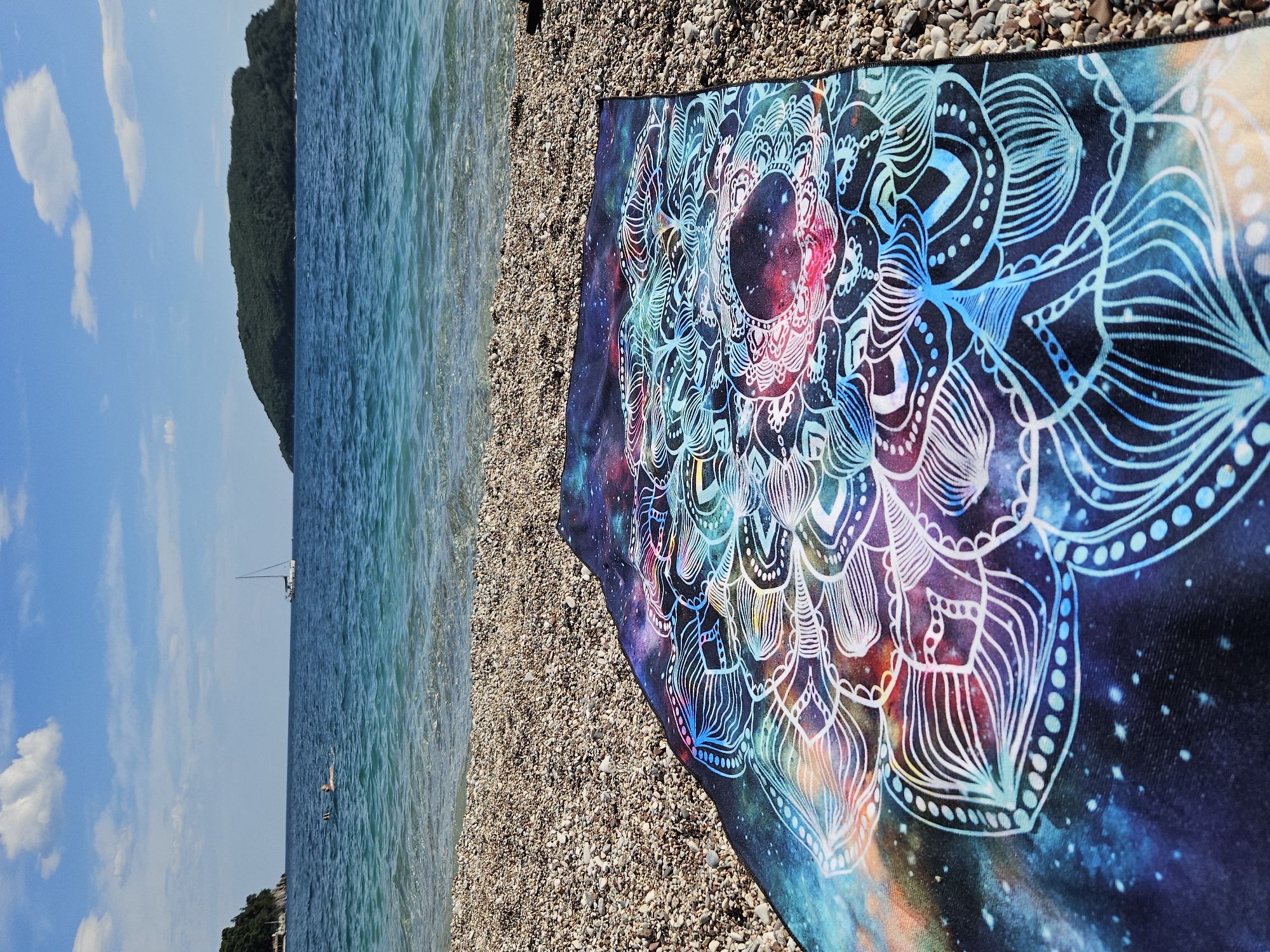 The image shows a beach scene, with a mandala-patterned towel laid out on a pebble-covered section of the beach. The ocean is visible on the left side of the frame, with a sailboat in the distance. The sky is blue with scattered clouds. A land mass with trees and vegetation can be seen on the left. The beach appears to be located in an unknown city and state.