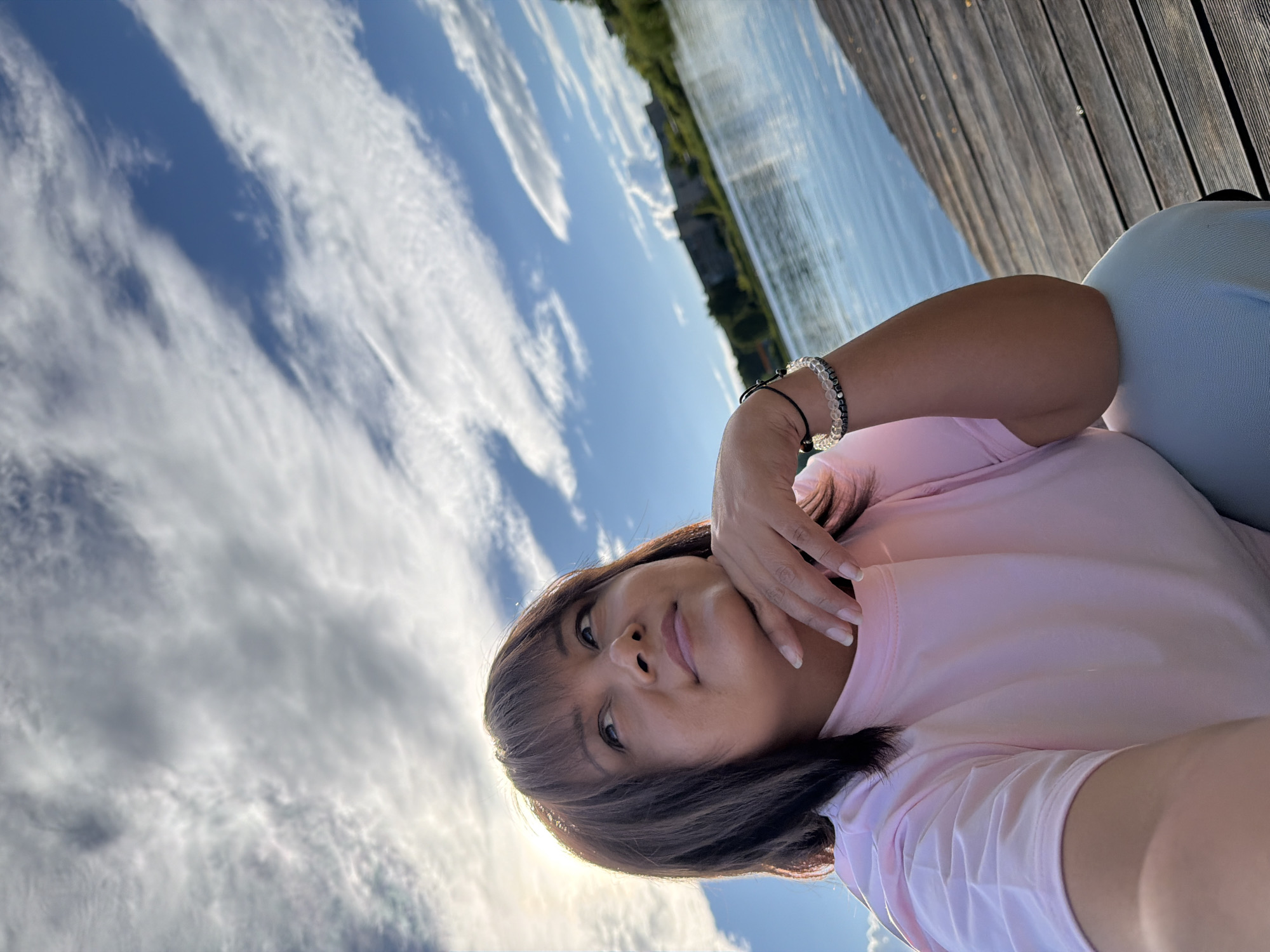 A medium shot captures a woman in a light pink t-shirt and light blue pants, looking up and to the left with her chin resting on her hand. She has dark brown hair and is wearing bracelets on her left wrist. The background shows a body of water with a wooden pier in the foreground, and a distant building or ruins on the far bank. The sky is a bright blue with scattered white clouds, and the sun is visible behind the woman's head, casting a lens flare. The image is taken from a low angle, looking up at the woman.