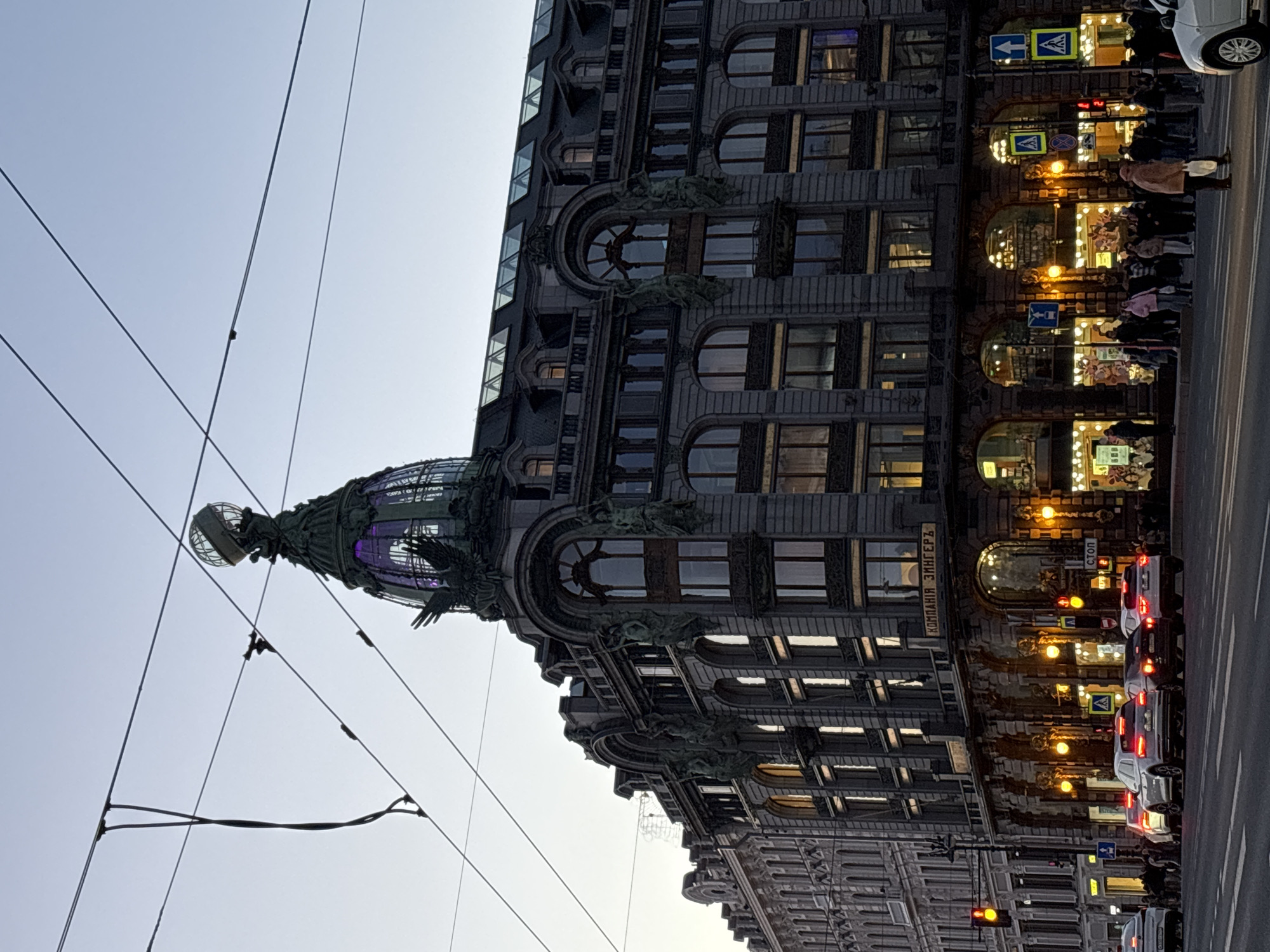 The image shows the Singer House, also known as the House of Books, located in Saint Petersburg, Russia. The building has a notable dome structure. Wires for public transportation are visible against the sky. The building's facade is detailed with ornate architectural elements. Pedestrians are seen walking along the sidewalk in front of the building. Vehicles, including cars, are present on the road next to the sidewalk. Street signs and traffic lights are also visible. The time of day appears to be dusk or early evening, as suggested by the ambient light and illuminated shop windows.