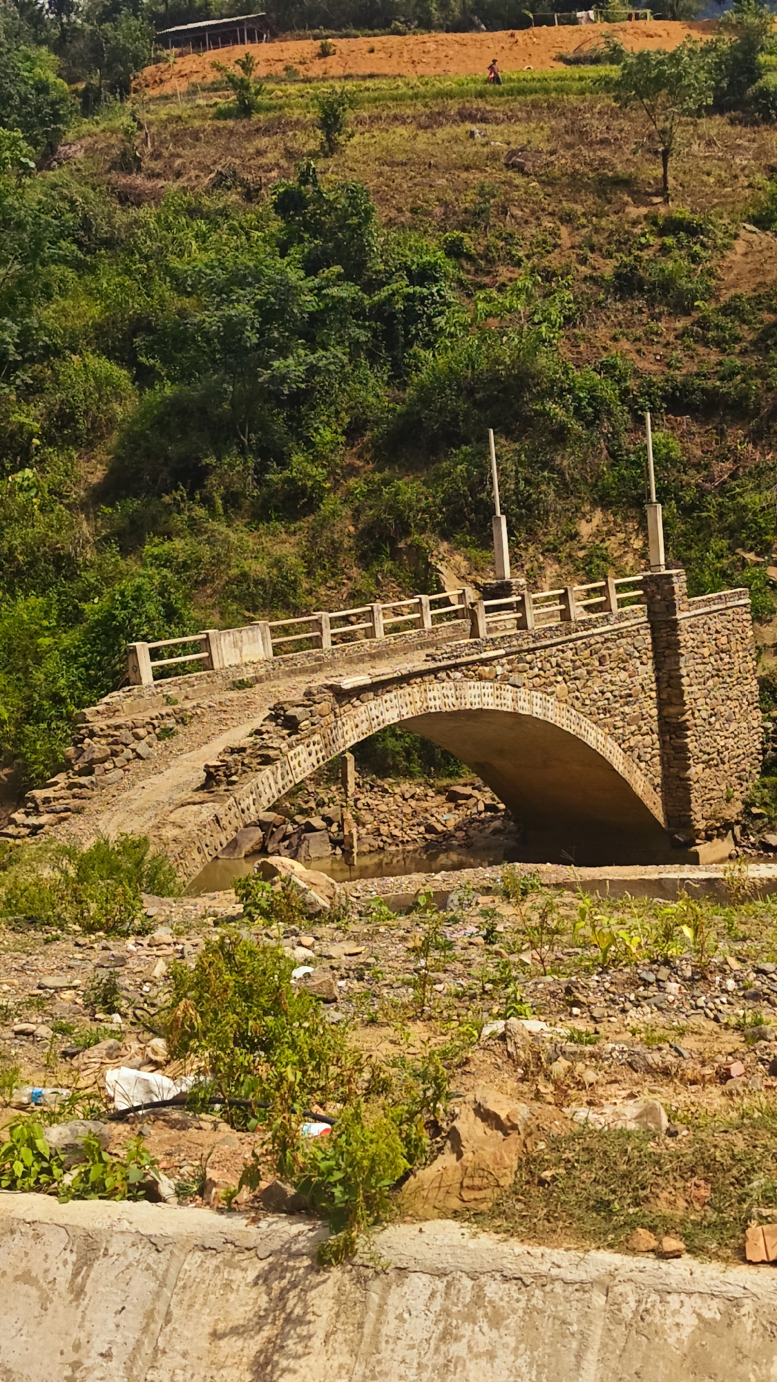 This image presents a vibrant rural landscape, centered around a rustic stone and concrete arch bridge in what appears to be Khao Mang, Vietnam. The bridge, the primary subject, is constructed with irregular stone masonry forming its arch and side walls, topped with a simple concrete railing. Two tall, white poles stand on the bridge deck, possibly for utility lines. Beneath the bridge, a narrow, shallow stream flows over a rocky bed.
The background is dominated by a steep hillside blanketed in lush green vegetation and dense trees. Several patches of exposed, reddish-brown earth are visible on the slope, suggesting agricultural activity or land clearing. On one of these higher, cleared sections, a small human figure, identifiable by a bright red garment, can be seen, likely engaged in farming. To the upper left, a basic, open-sided structure with a dark roof is nestled against the hillside.
The foreground consists of rocky, earthy terrain with sparse green plants. Some debris, including plastic waste and bricks, is noticeable near the bottom edge of the frame. The scene is brightly lit, indicating a clear, sunny day, likely morning or afternoon. The overall impression is one of natural beauty intertwined with human infrastructure and rural life in a hilly region. No visible text is present in the image.
