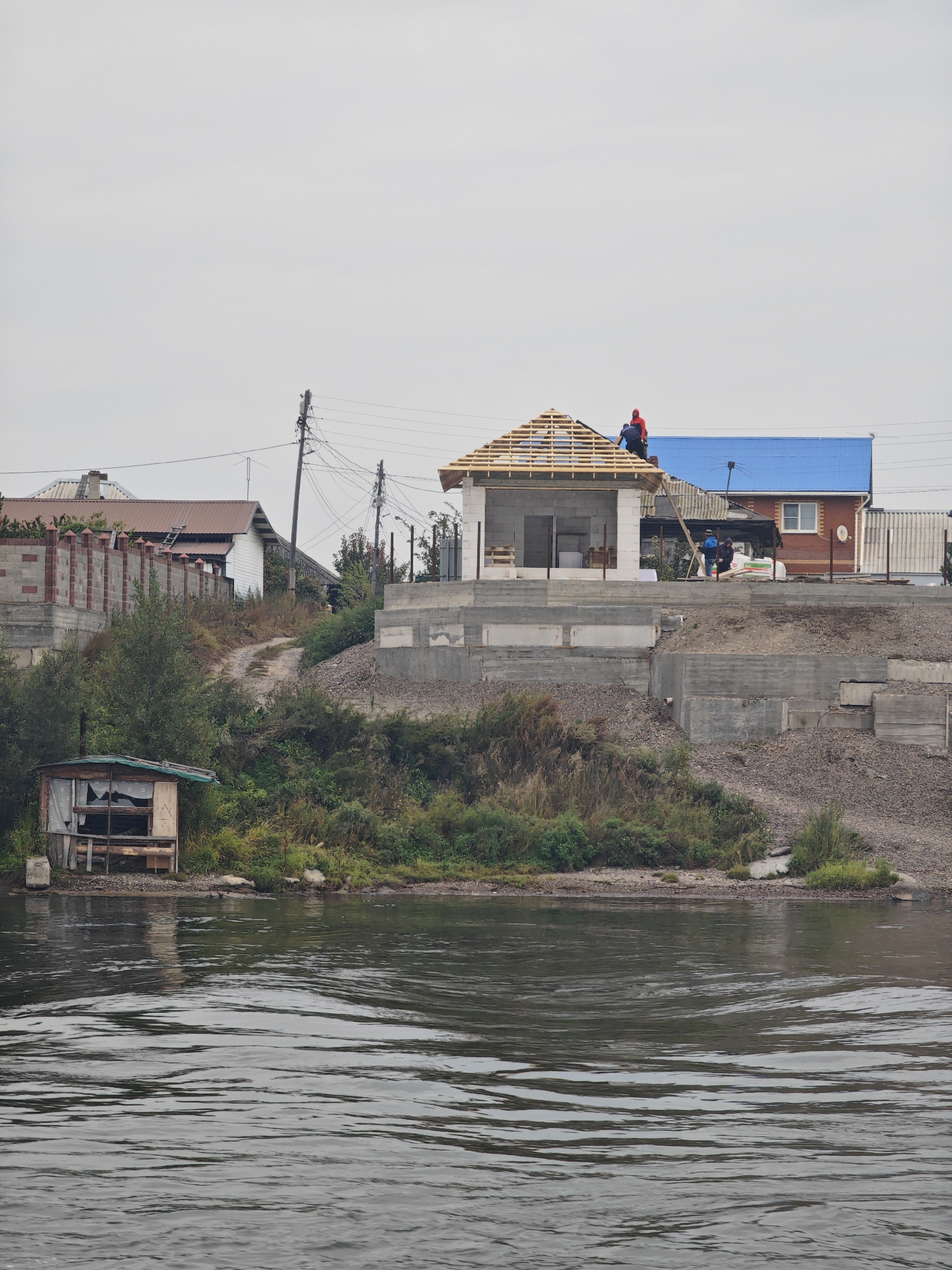 The image depicts a construction site on the bank of a river in Ermolaevo, Russia, under an overcast sky. In the foreground, the rippling surface of the dark river occupies the lower half of the frame. The middle ground features a vegetated embankment leading up to several structures. On the left, a small, dilapidated wooden shed with a green roof and broken windows sits on the water's edge. Further back and uphill, a road winds between dense green bushes and trees.
The upper portion of the image showcases a residential area under construction. A house with a wooden roof frame is being worked on, with two individuals on the roof, one in a red hoodie and another in a blue shirt, engaged in construction activities. To the right of this house is a finished building with a bright blue metal roof. Behind these, other houses with various roof types and some construction materials are visible. The ground level of the construction area is characterized by concrete foundations and retaining walls. Electrical poles and wires are present, indicating the presence of infrastructure. The overall impression is one of ongoing development in a semi-rural or suburban setting.