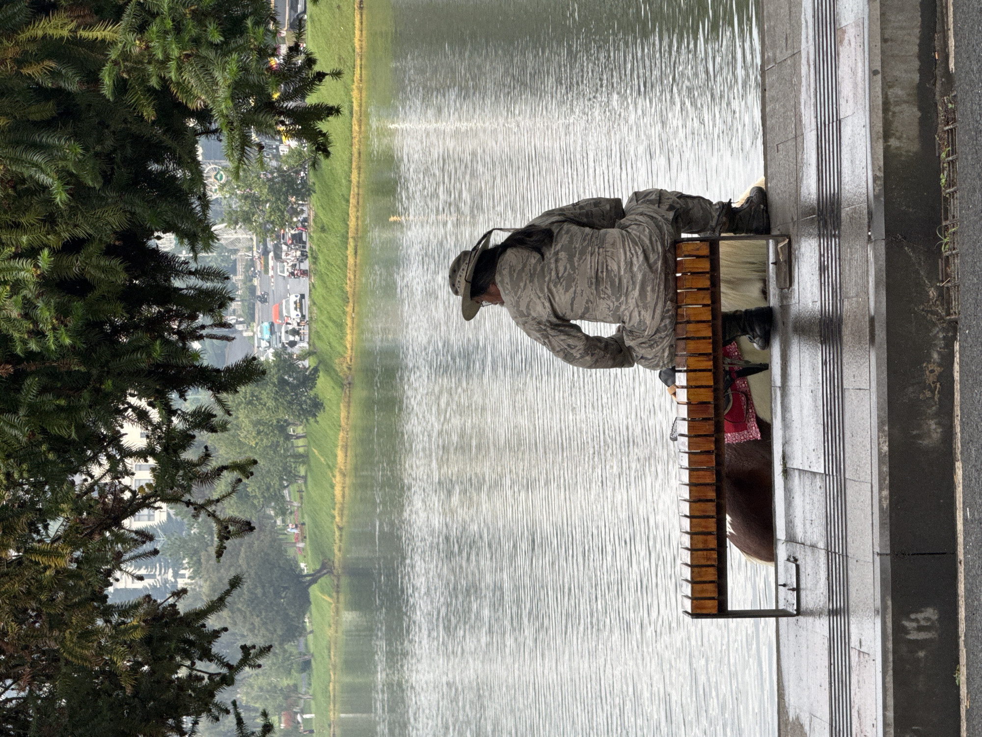 A person wearing a camouflage uniform and a wide-brimmed hat is sitting on a wooden bench facing a large body of water. The bench is positioned on a paved walkway next to the water. Behind the person, a portion of a horse, covered with a reddish blanket, is visible. The horse appears to be standing in the water or at its edge. The water is calm with ripples, reflecting the overcast sky. Trees with lush green foliage overhang the scene from above, casting shadows. In the background, across the water, a city landscape is visible with buildings and trees. A road with some vehicles can also be seen in the distance, suggesting an urban park setting. This scene is located at Xuan Huong Lake in Da Lat, Vietnam, a popular tourist destination known for its natural beauty and recreational activities. The overall atmosphere is serene and contemplative.