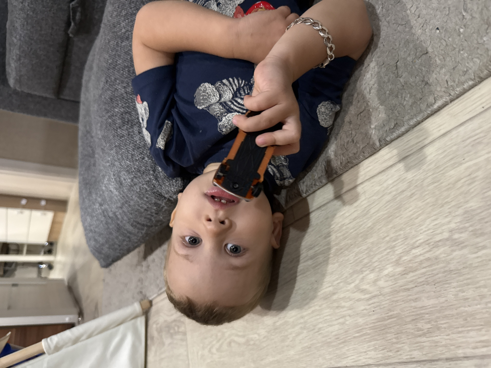 This media file features a young child lying on their back on a wooden floor with a rug. The child is wearing a dark blue t-shirt with a tiger pattern and a silver bracelet on their left wrist. They are holding a small orange and black toy car in their hands, bringing it close to their mouth. The child has blue eyes and light brown hair.
The scene appears to be indoors, possibly a living room or play area, with a grey sofa visible in the background and a light-colored rug partially covering the wooden floor. The lighting suggests it is daytime, and there are no obvious indications of specific weather conditions or a particular time of day. The child's expression is one of curiosity or playfulness. There is no visible text within the image. The overall setting is casual and domestic.