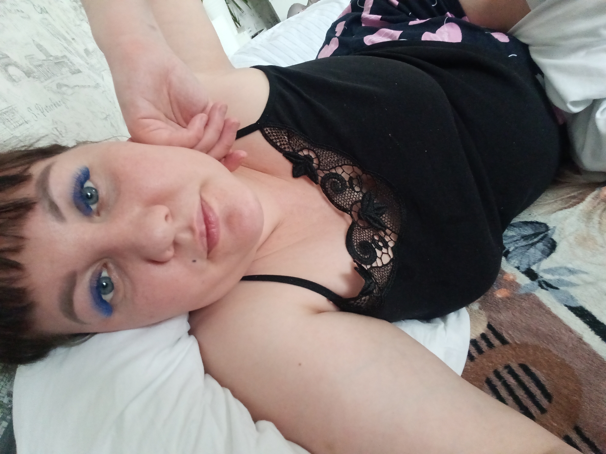 A woman is lying on a bed, smiling and looking at the camera. She is wearing a black tank top with lace trim and black shorts with pink hearts. She has blue eyeshadow on her eyelids. The bed has a white pillow and a patterned blanket. The location is not identifiable as Osa, Russia based on the image.