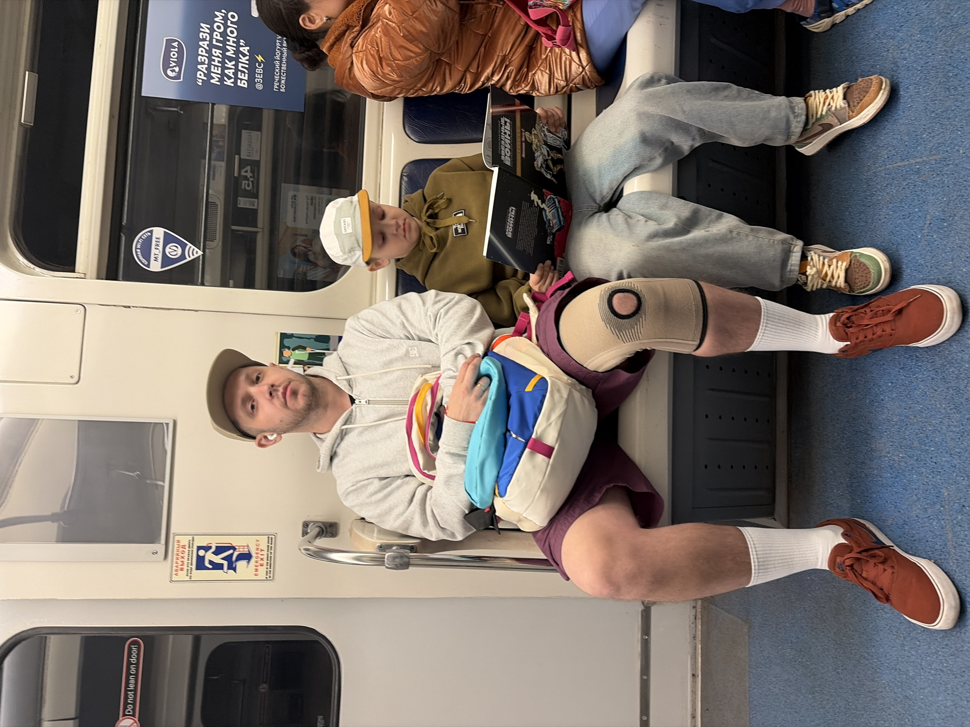 The image shows the interior of a subway car in Saint Petersburg, Russia. Three individuals are seated on a row of seats. The individual closest to the camera is a light-skinned man with short hair, wearing a light gray hooded sweatshirt, maroon shorts, white socks, and rust-colored sneakers. He has a tan baseball cap and is wearing an earbud in his left ear. He carries a colorful backpack. He has a beige-colored knee brace on his right knee. Next to him sits a younger, fair-skinned person wearing a tan hoodie, a white baseball cap with a yellow bill, light gray denim jeans, and tan and brown sneakers with white shoelaces. This person is holding two books. Next to them is an older person wearing a brown puffy coat, faded light gray denim jeans, and tan and brown sneakers with white shoelaces. The walls of the subway car are a mix of white and gray, with a handrail visible in front of the man. Various signs and stickers are present on the windows and walls of the subway car. These include safety instructions for emergency exits and a sticker indicating free Wi-Fi. There is also an advertisement in Russian for yogurt. The floor is blue.