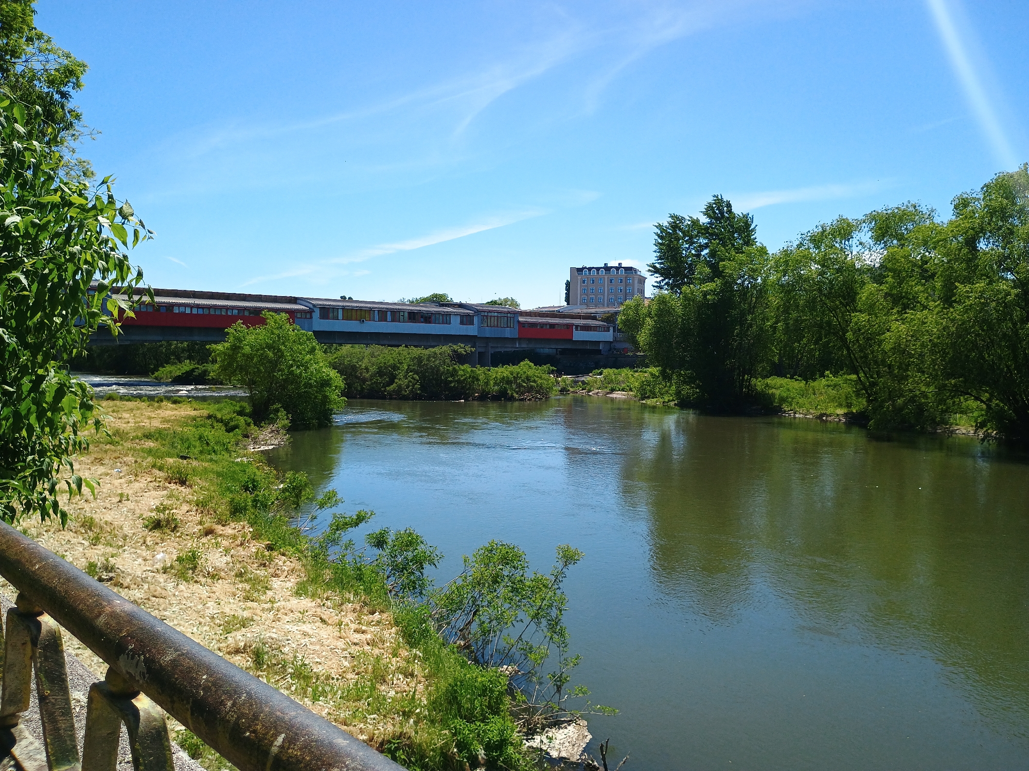 The image shows a view of a river with trees lining the banks. A multi-car train or tram is on a bridge over the river, with a building visible in the background. A rusty metal railing is in the foreground, partially obscuring the view of the landscape. The sky is blue with some light clouds.