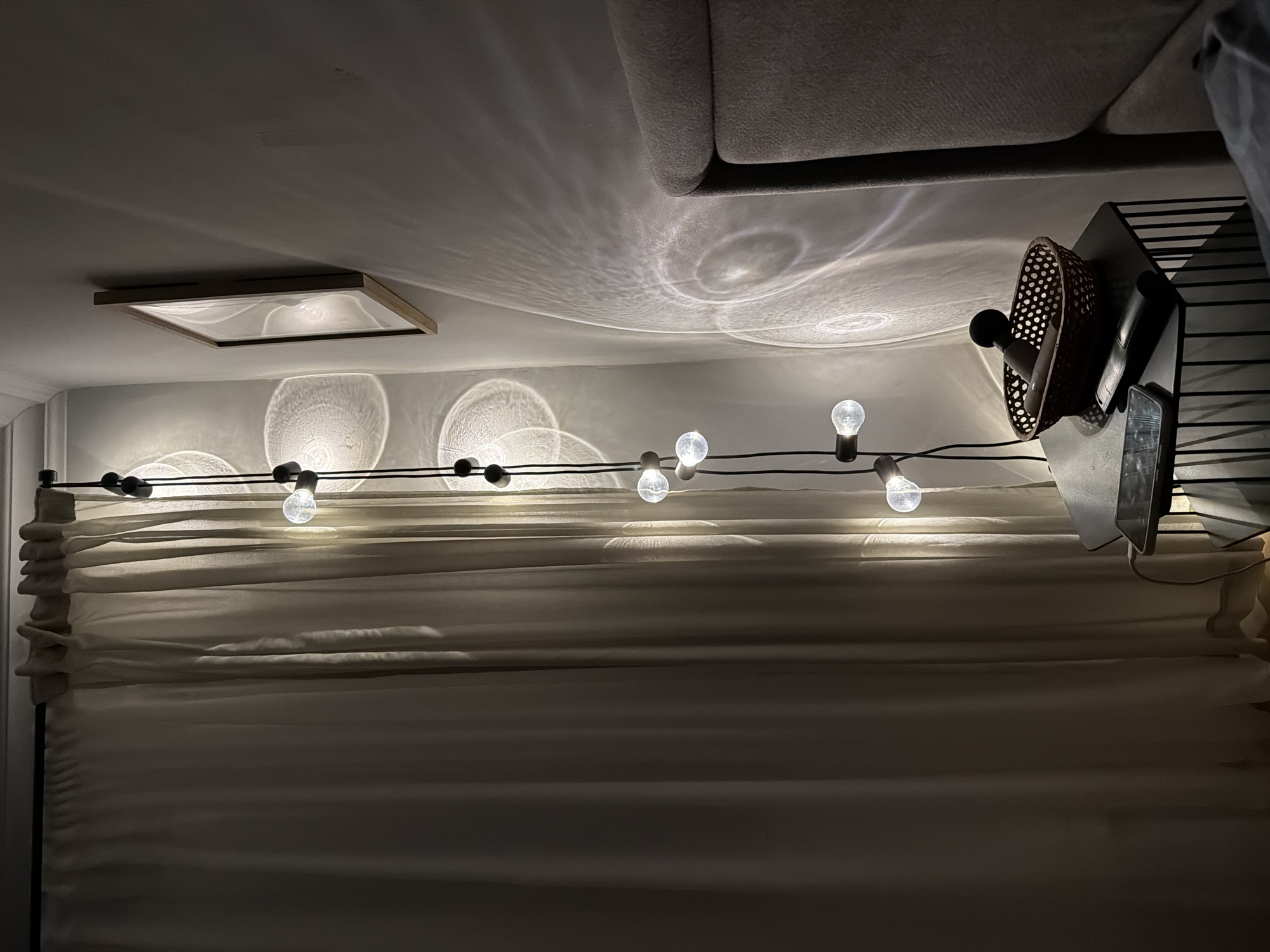 The photograph shows a dimly lit interior, likely a room in a residential space in Khimki, Russia. A string of lights with clear, round bulbs is draped horizontally across the frame, casting circular patterns of light onto the ceiling and wall. Below the lights, a light-colored fabric curtain or drape with folds is visible. In the upper-right corner, part of a gray upholstered couch is discernible, with light reflecting from the ceiling. A modern-style light fixture is inset into the ceiling to the left. A dark-colored shelving unit or rack is visible on the right, with objects, including a round wicker basket and a rectangular device, sitting on it. The overall impression is of a room illuminated by the decorative string lights.