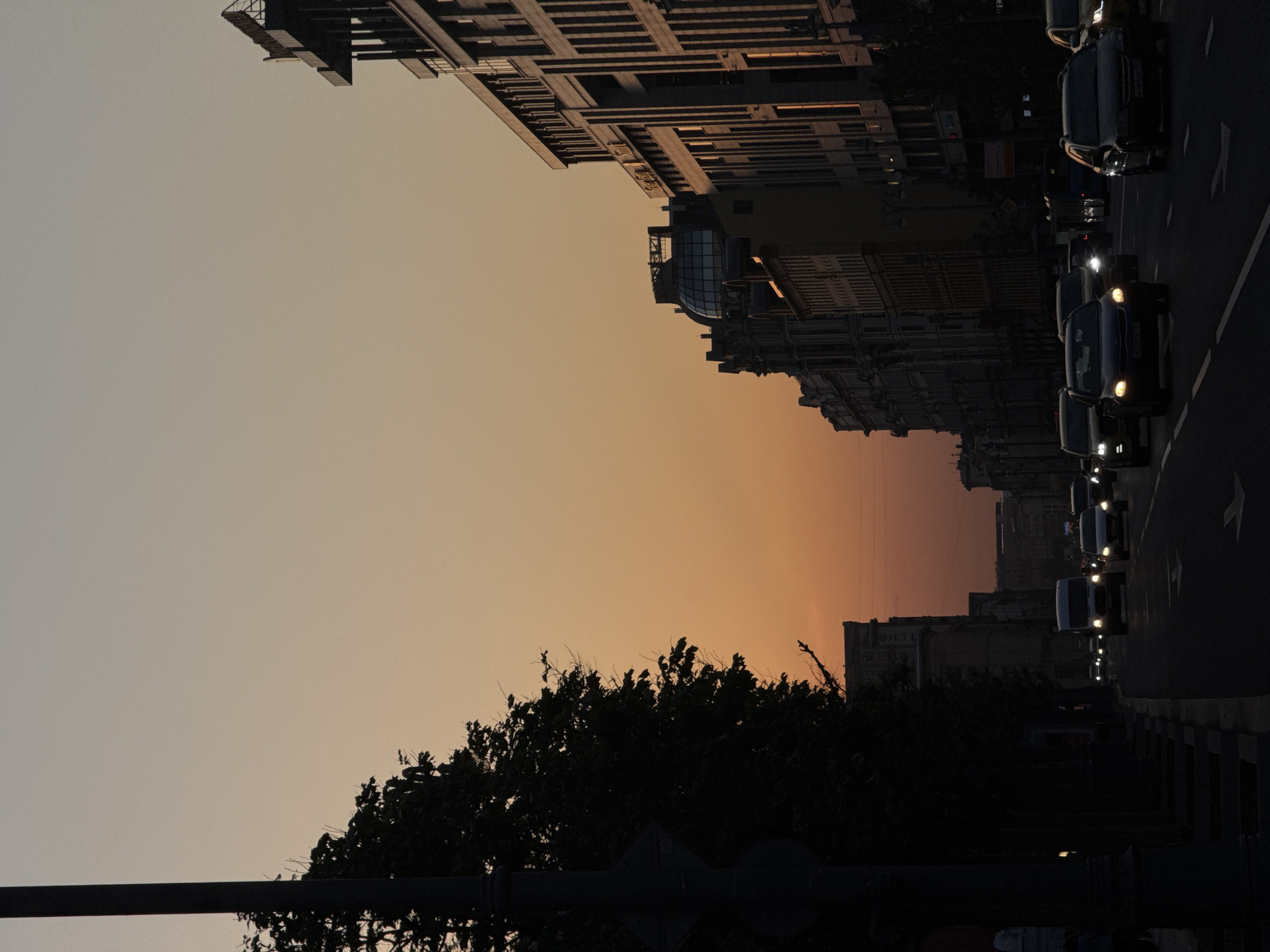 The image depicts a Moscow street at sunset. The perspective is from ground level, looking down a road lined with buildings. The sky transitions from a pale orange at the horizon to a lighter shade above. On the right side of the image, cars with headlights on are traveling down the street. On either side of the street are buildings, with one building featuring a glass dome structure. Trees are silhouetted in the lower portion of the frame, and a street pole is visible in the foreground.