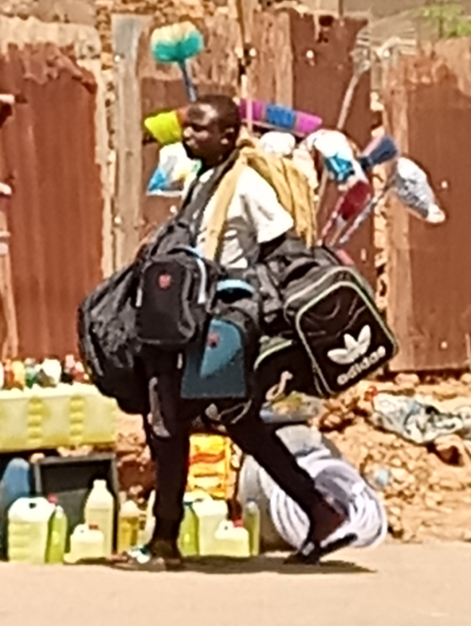 In an outdoor setting within the city of Maiburiji, Nigeria, a male figure is seen carrying multiple bags and items. He has on a white shirt and dark pants. A black Adidas bag is visible. The items being carried include what appear to be cleaning supplies, such as brooms, and what appear to be small canisters of cooking oil. The background appears to be a somewhat cluttered and disorganized urban environment.