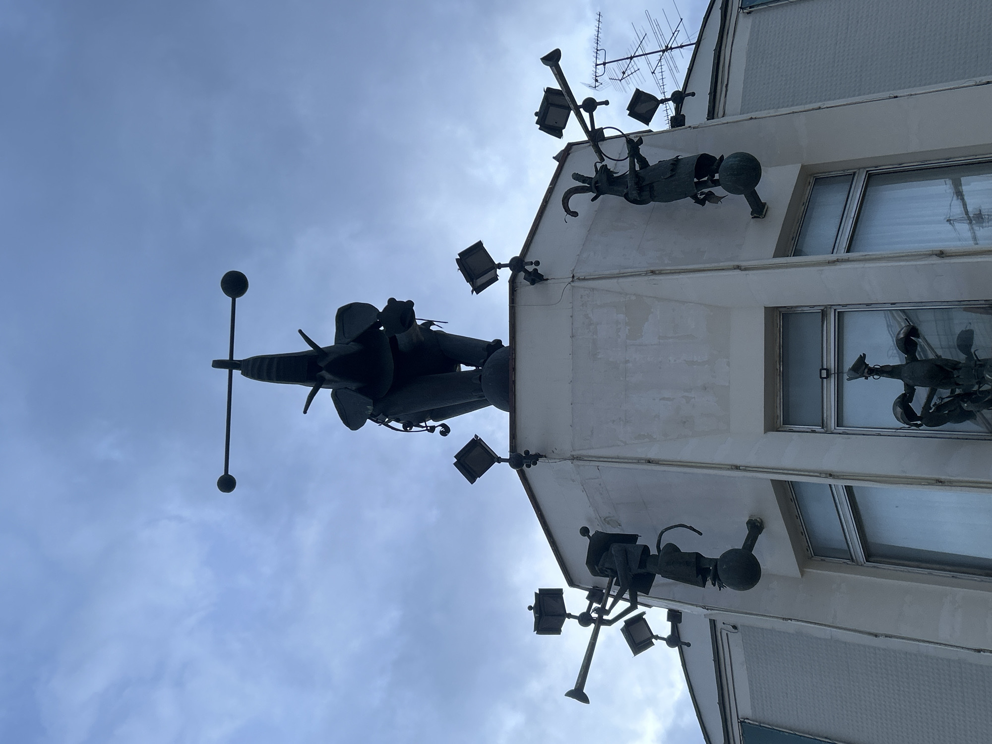The image, taken from a low angle looking upwards, displays the upper section of an off-white building against an overcast, light blue-grey sky. The building's facade is heavily adorned with dark, metallic sculptures that extend outwards, appearing as stylized or abstract figures, possibly human or mythical. These sculptural elements are integrated into the architecture. Several dark light fixtures or spotlights are also mounted on the building's exterior and among the sculptures.
On the rooftop, multiple antennae are visible, including a prominent, complex TV aerial array. A tall, slender pole with a spherical top and a horizontal crossbar, resembling a weather vane or an architectural finial, stands silhouetted against the sky from the highest point of the structure.
Windows on the building reflect the cloudy sky, and the overall lighting is soft and diffused, characteristic of an overcast day. No text is visible in the image. The scene captures an urban architectural detail with artistic embellishments.
