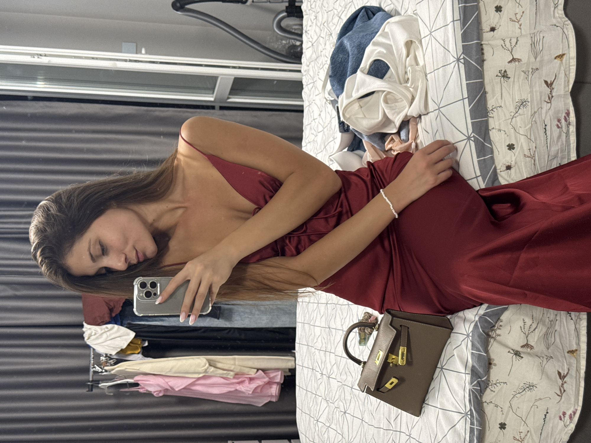 A woman in a dark red slip dress is taking a selfie in a bedroom. She has long brown hair and is wearing a white beaded bracelet. In front of her, on the bed, is a small brown handbag with gold hardware. The bed is covered with a white and grey patterned duvet. Behind her, clothes hang on a rack, and a vacuum cleaner is visible against the wall. The scene is likely indoors, possibly a residential setting. The mention of Burgas, Bulgaria, is not supported by visual evidence in the image.