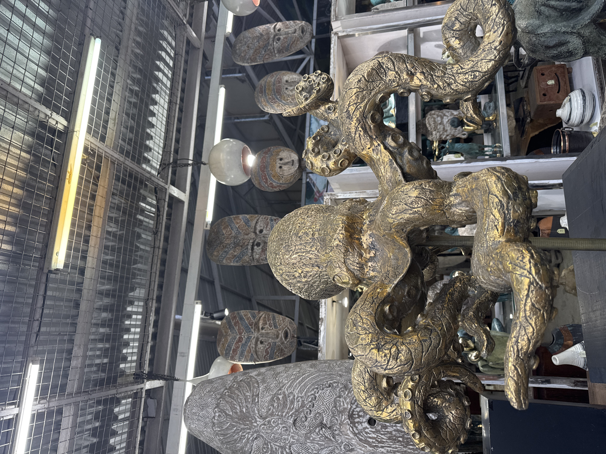 The image shows an indoor space, possibly a shop or a display area, with a variety of handcrafted items. The focus is on a complex, golden-colored, sculpture. Behind the sculpture, several flat masks with various patterns are displayed, appearing to hang from the ceiling. The ceiling area is a structure of metal grids and fluorescent tube lights. The image was taken in Ubud District, Indonesia.