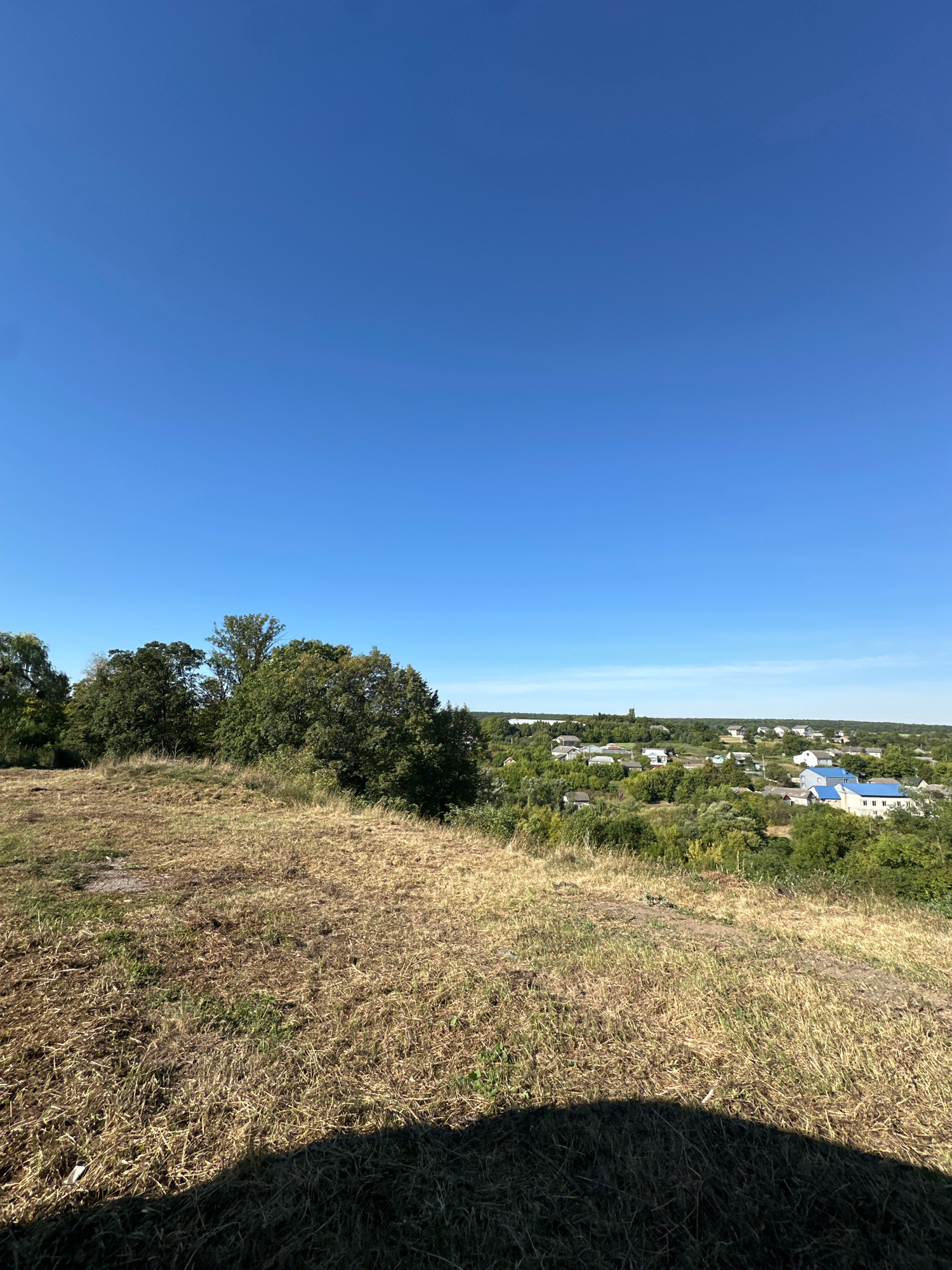 This is a photograph of a rural landscape under a clear blue sky. In the foreground, a grassy hill slopes downwards, covered in dry, straw-colored grass. The middle ground features a dense line of trees, with varying shades of green foliage. Beyond the trees, a village is visible, characterized by scattered houses with predominantly white and blue roofs. The architecture appears to be typical of a residential area in the Ukrainian countryside. The overall impression is one of a serene, sunny day in a peaceful rural setting. This appears to be a view from an elevated position overlooking the village of Makiv, Ukraine.