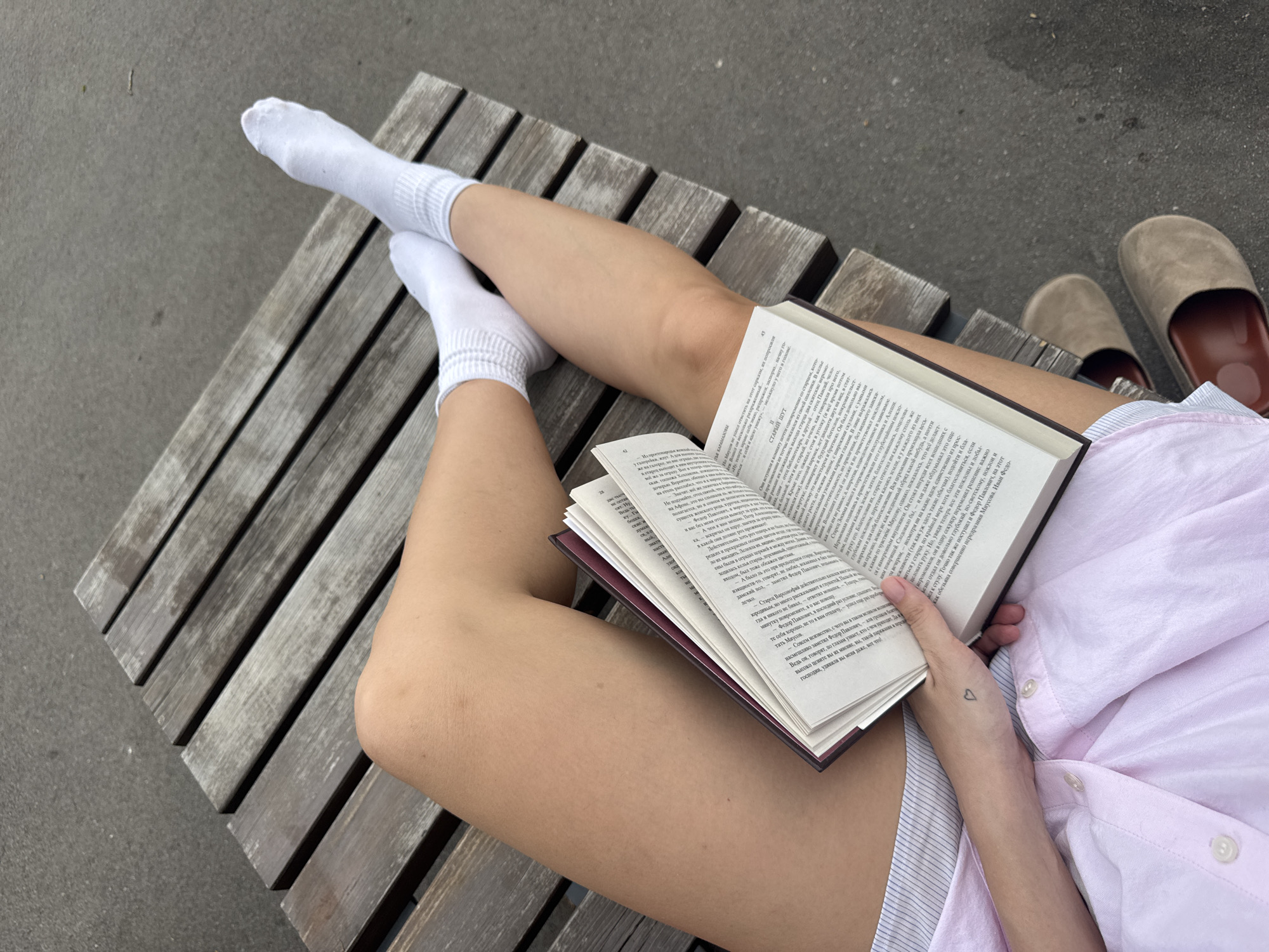 A young woman is seated on a wooden bench outdoors, likely in a public area. She is wearing white socks and a light pink, unbuttoned shirt. Her legs are crossed, with a book resting on her thigh. The book is open, revealing dense text in what appears to be Russian. She is holding the book open with her left hand. A small heart tattoo is visible on her left wrist. A pair of brown slippers is visible on the right side of the frame. The bench is made of weathered wood slats. The ground is a plain, gray concrete surface. There is no other information that would place this specifically in the city of Saratov, Russia.