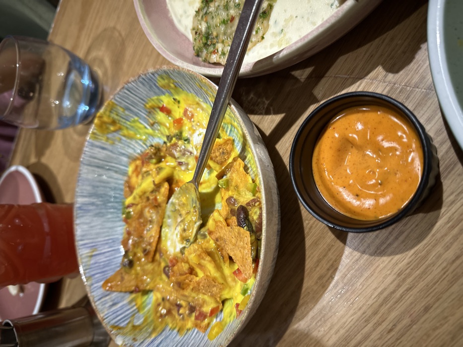 The image displays an indoor dining table scene. In the foreground, a black, ribbed ramekin holds a vibrant orange-red dipping sauce. To its immediate right, a portion of a light-colored bowl is visible. Dominating the midground is a partially consumed dish of nachos in a ceramic bowl with a blue and white striped rim. The nachos consist of tortilla chips coated in melted yellow cheese, black beans, diced red peppers, and green herbs. A silver spoon rests in the nacho mixture, extending towards the right. To the right of the nacho bowl, a pink-rimmed bowl contains a white, creamy dish topped with green herbs and red flakes. Behind this, a clear glass with a blue-tinted base is present. In the background to the left, a red beverage in a textured glass and a silver condiment shaker are visible, alongside a blurred pink plate. The entire scene is set on a light brown wooden table exhibiting a linear grain pattern. No specific architectural details or signage are visible to determine the exact location within Kalyan-Dombivli, India, beyond being an indoor restaurant or cafe setting. No individuals are fully visible, though human presence is implied by the dining arrangement.