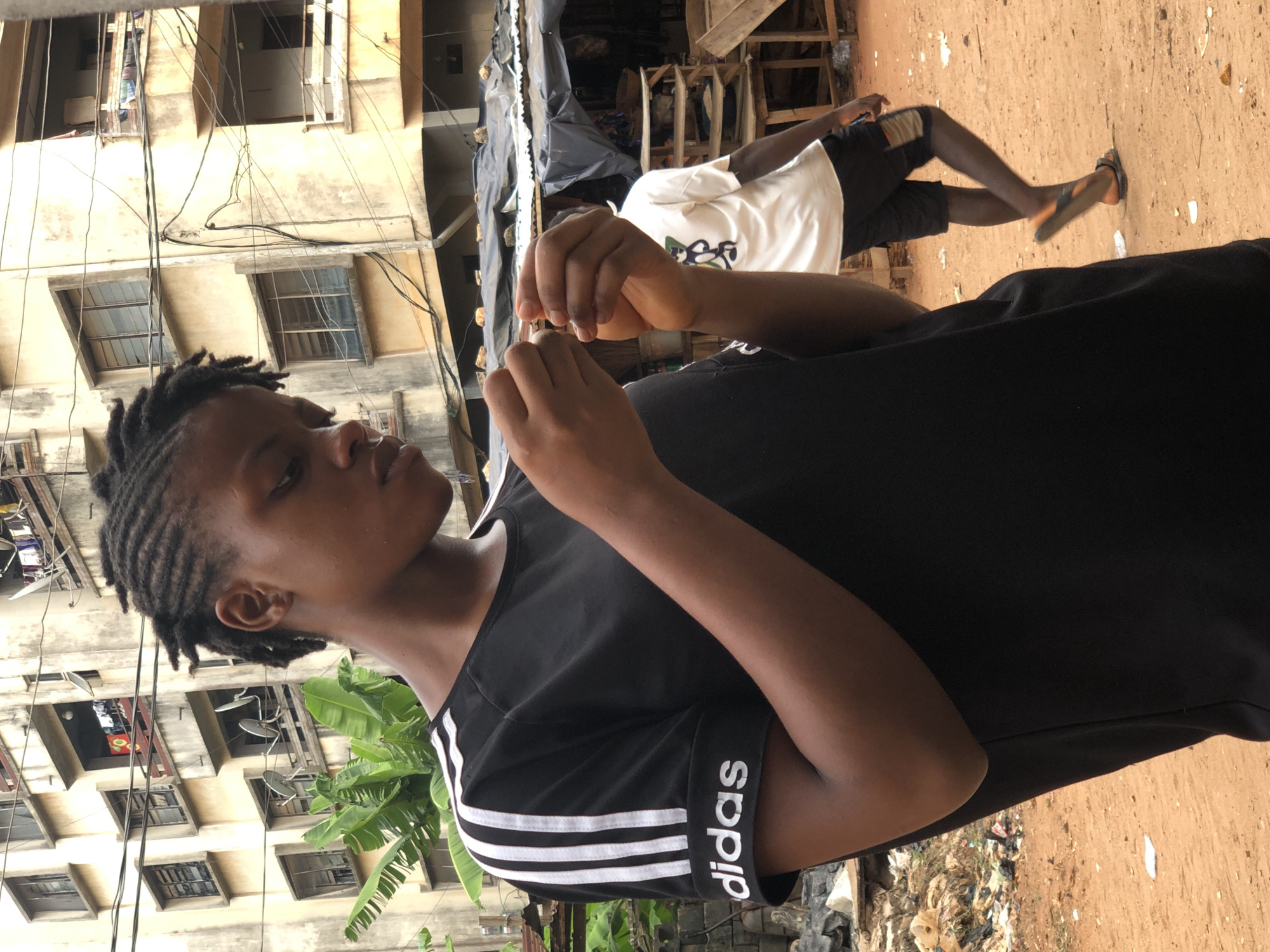 The image is rotated 90 degrees clockwise, presenting an outdoor scene dominated by two individuals. In the foreground, a young person with dark skin and braided hair is featured from the chest up. They are wearing a black short-sleeved t-shirt with three white stripes on the sleeves and the word "adidas" printed vertically in white on the right sleeve. Their hands are raised and positioned in front of them, holding and seemingly manipulating a small, light-colored object with focused attention.
Behind and slightly to the right of the main subject, a second person with dark skin is visible from the waist down, wearing a white t-shirt, black shorts, and flip-flops. This person appears to be walking away from the viewer, moving towards the right side of the frame.
The background reveals a multi-story, light-colored building, possibly concrete or stucco, with numerous windows. A complex network of dark wires traverses the building's facade. To the right of the main subject, large green leaves, possibly from a banana plant, are visible. The ground is an unpaved, reddish-brown dirt surface, with scattered debris and trash evident towards the lower right. The scene is brightly lit, suggesting clear, sunny daytime conditions in what appears to be an urban or semi-urban environment.