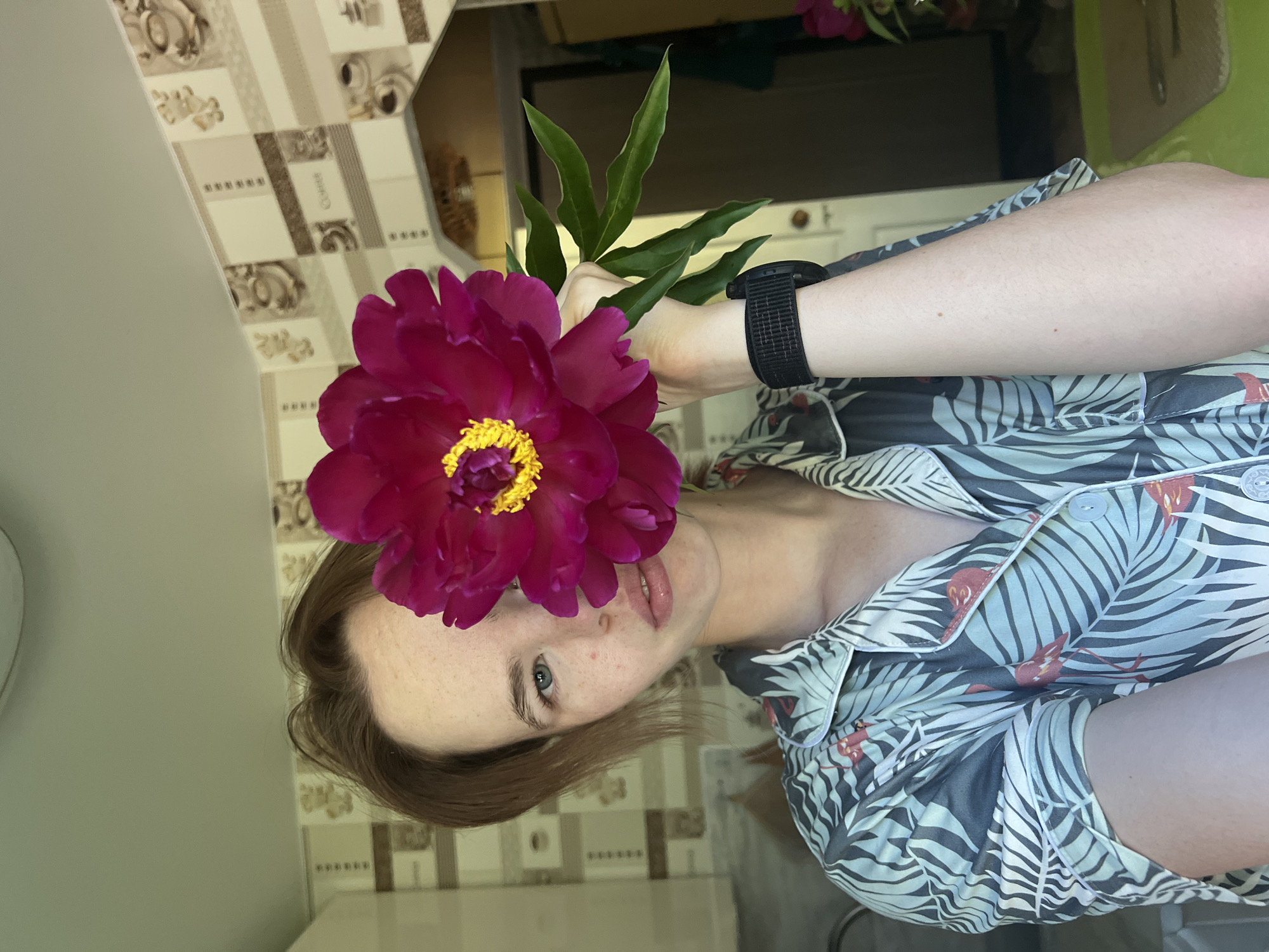 A person stands indoors, holding a large, dark pink peony flower in front of their face, obscuring their right eye and nose. The person has light brown hair and is wearing a short-sleeved shirt with a tropical print featuring palm leaves and flamingos. The background shows a kitchen area with patterned wallpaper depicting coffee cups and mugs. The environment appears to be an apartment in Chaikovsky, Russia. The lighting suggests it is daytime.