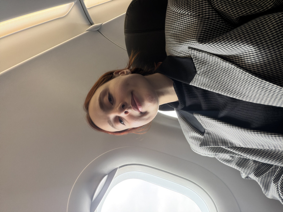 The image depicts a female individual inside an aircraft cabin, consistent with a flight associated with Tuzla, Turkey. She has reddish-brown hair and is wearing a black ribbed turtleneck under a grey-and-white patterned blazer with black lapels. The individual is positioned centrally, looking directly at the camera with a slight smile, exhibiting visible dimples. To her left, an oval airplane window reveals a bright, undifferentiated sky, indicative of daylight. The aircraft's interior features light-colored overhead panels and integrated strip lighting. A dark-colored seat headrest is partially visible behind the individual.