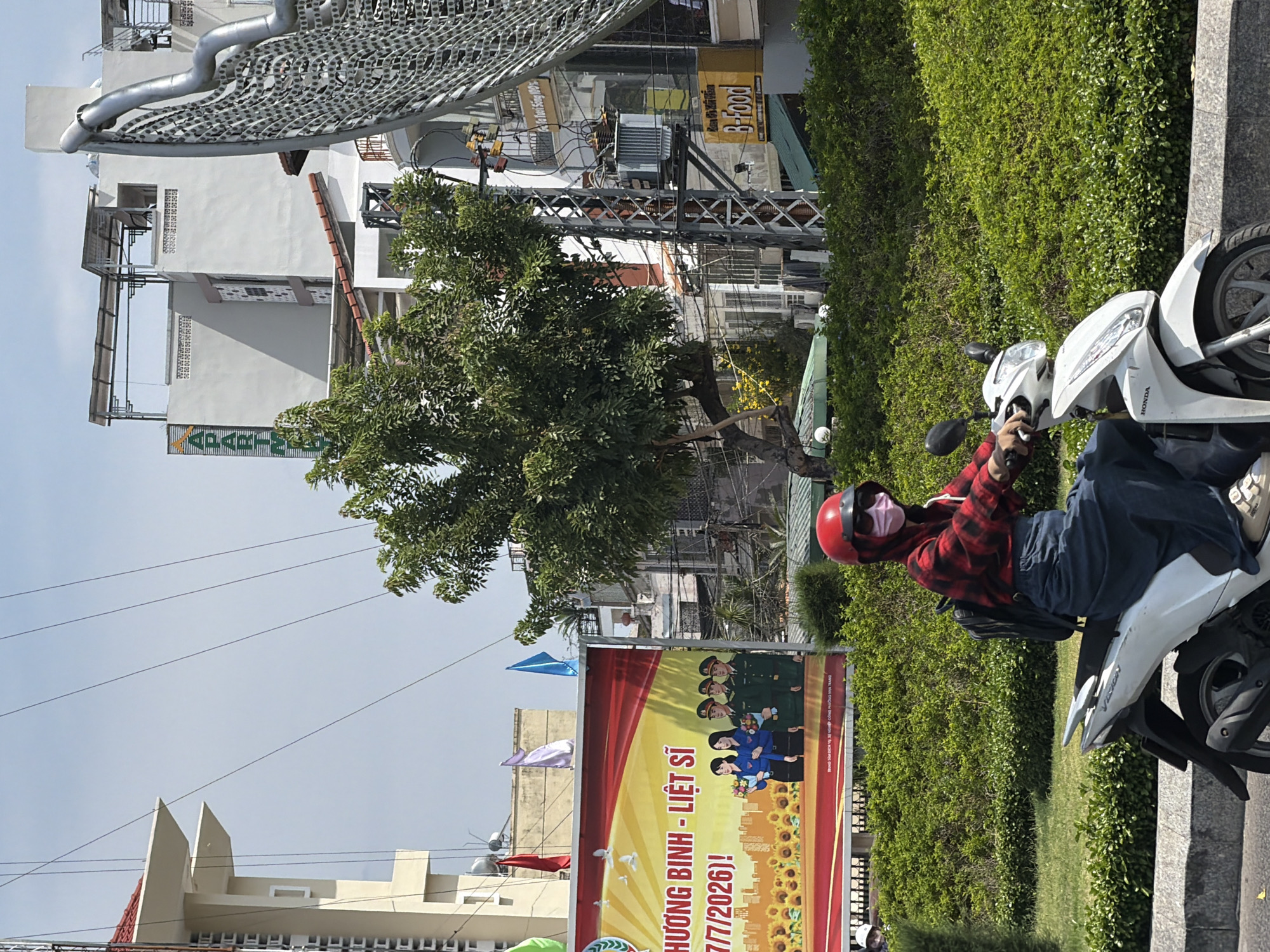 A person wearing a red helmet, sunglasses, and a pink face mask rides a white Honda scooter on a sunny day. They are wearing a red and black checkered shirt and a long blue skirt, and a backpack is visible. The scooter is moving forward on a paved road with a grey curb. Behind the scooter, a densely planted green hedge forms a backdrop. In the background, a large yellow billboard displays images of sunflowers and people in military uniforms, with Vietnamese text. Further in the background, various buildings of different architectural styles, including a multi-story apartment building with the word "APARTMENT" visible, and utility poles with electrical transformers and wires are present. The sky is clear and blue. The scene appears to be in Nha Trang, Vietnam, based on the visual cues and the Vietnamese text on the billboard.