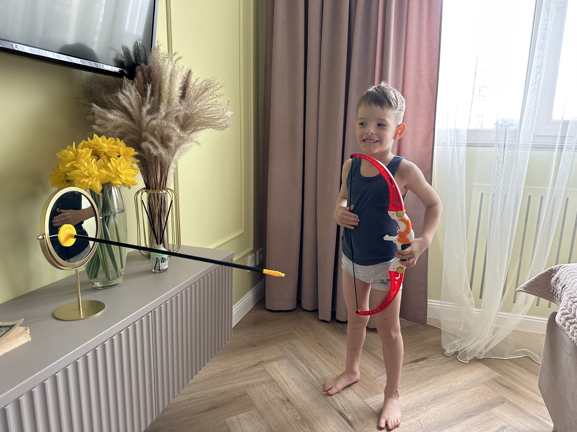 A young boy stands barefoot on a herringbone-patterned wood floor, holding a red and white toy bow. An arrow, black with a yellow tip, is aimed towards a table where a mirror reflects the arrow's trajectory. The background includes a gray cabinet, yellow flowers, and pampas grass. Behind the boy, there are pink curtains and a window covered with sheer white curtains. The scene appears to be indoors, possibly in a living room. The location is identified as Chaikovsky, Russia.