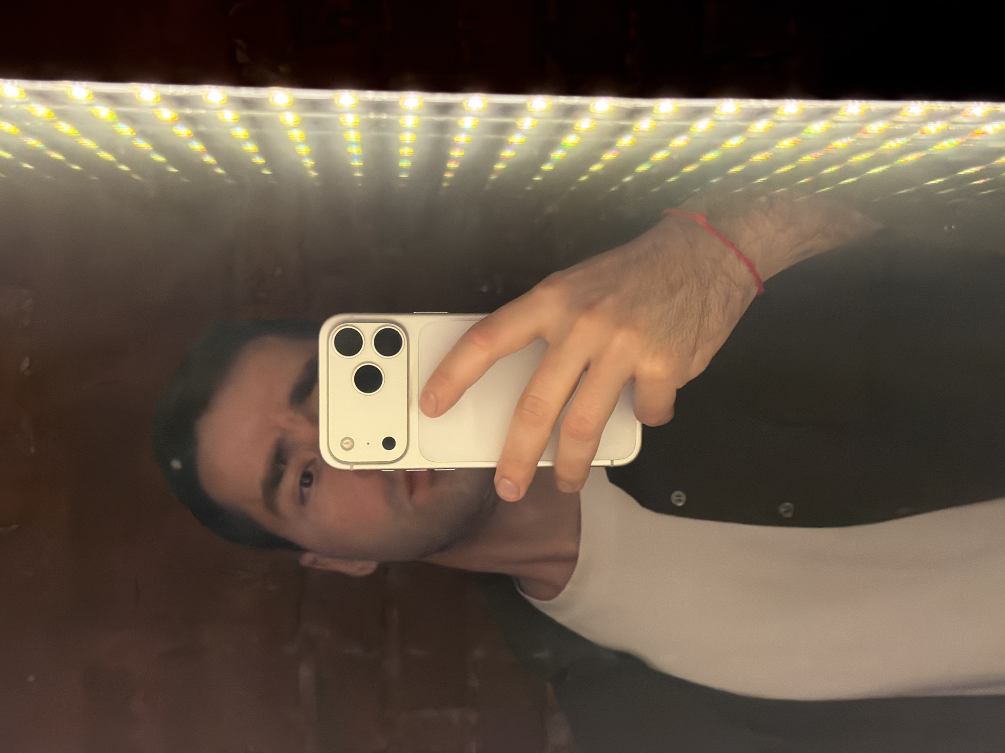 A man is taking a selfie in a mirror. The top of the mirror has a strip of bright LED lights. The man is holding a white smartphone. He is wearing a white shirt and a dark jacket. Part of a brick wall is visible in the reflection. There is no evidence to suggest the location is Moscow, Russia.