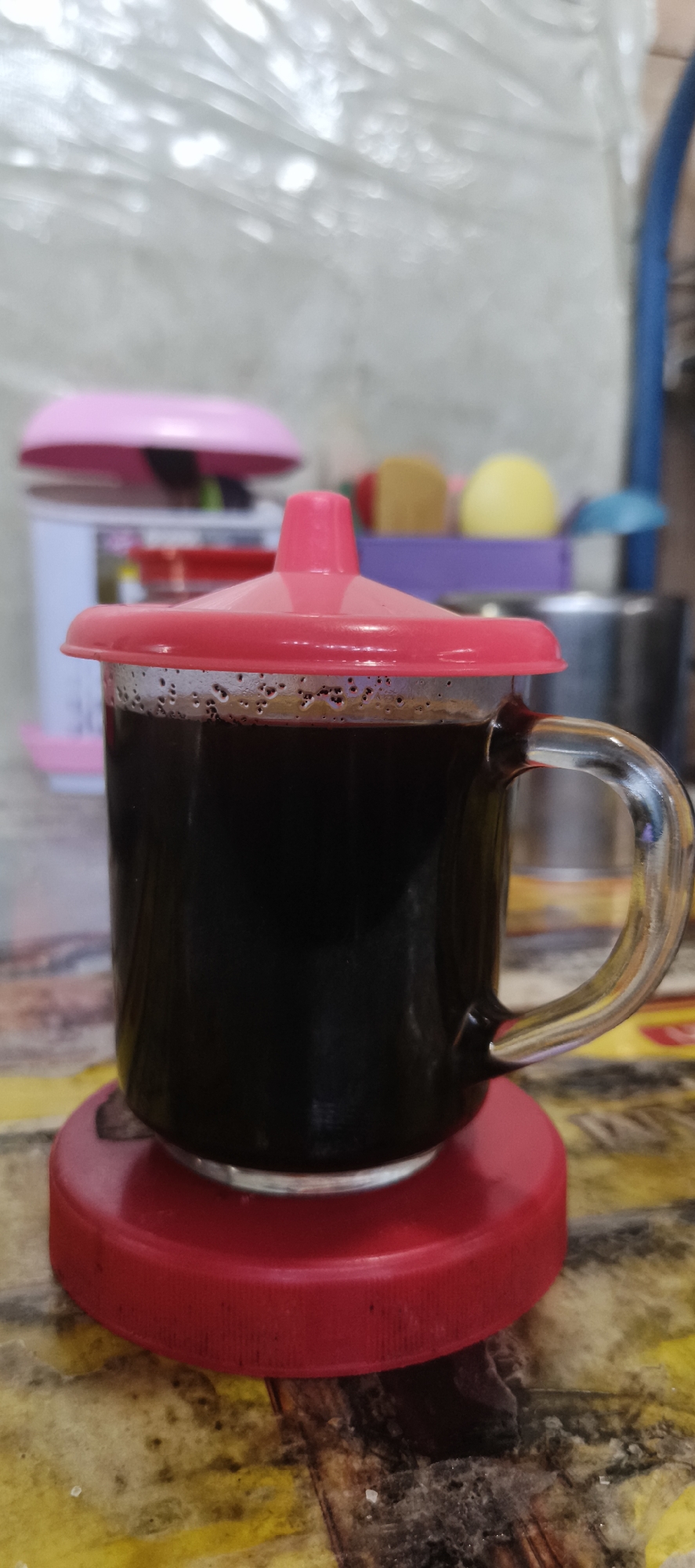 A close-up, eye-level shot shows a clear glass mug filled with dark liquid, likely coffee, topped with a bright pink lid. The mug is positioned on a red circular coaster, which in turn rests on a textured surface with yellow and brown tones. In the background, out of focus, are various household items, including a pink lid and a blurry pink and white object, suggesting an indoor setting, possibly a kitchen or dining area in a residence in Muaro Jambi, Indonesia. The lighting is soft, creating a slightly diffused effect.