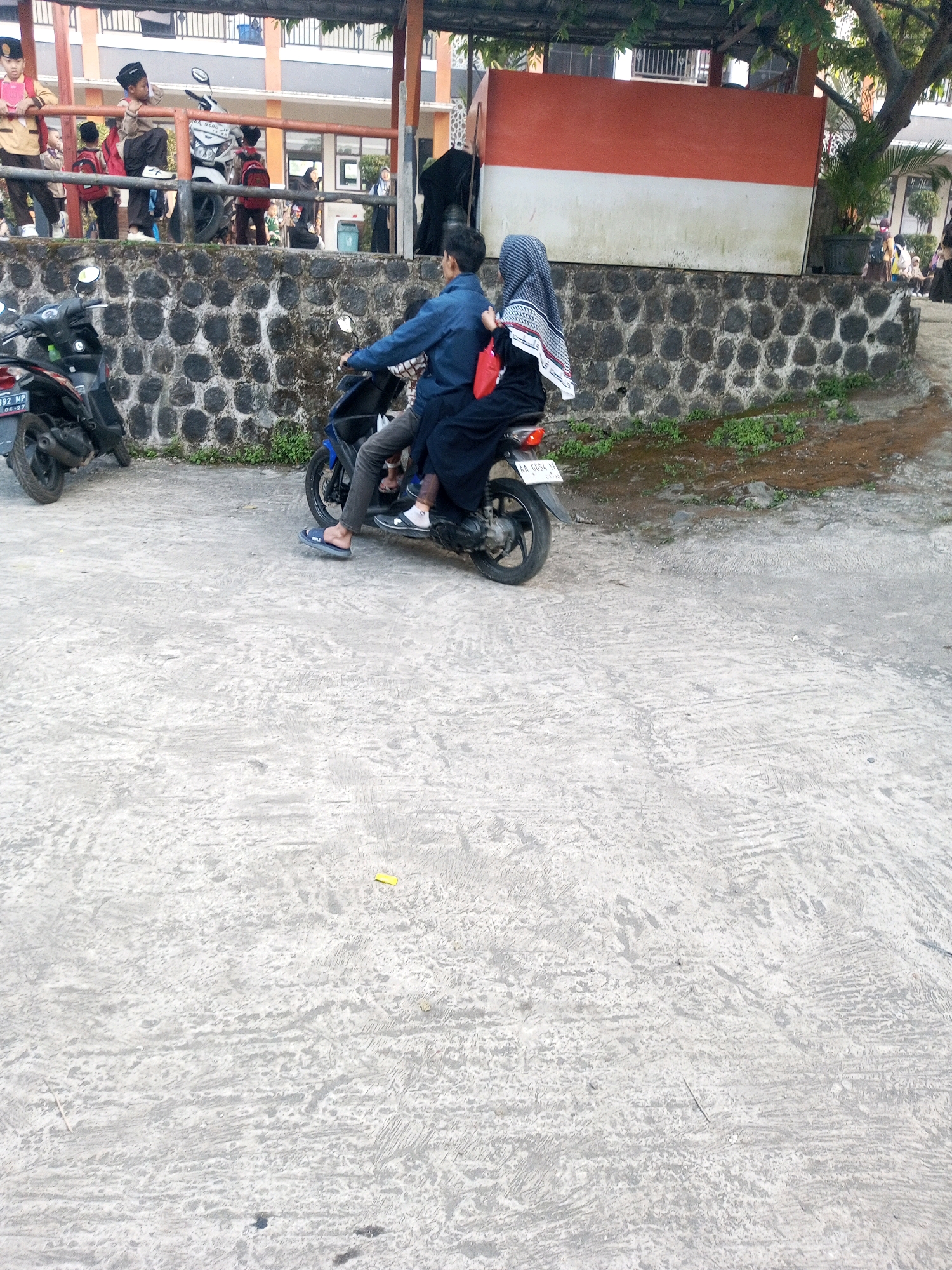 In Kalibeber, Indonesia, a man in a blue shirt and dark pants rides a scooter, carrying a woman behind him who wears a black dress and hijab. They are on a paved surface near a low stone wall. Behind the wall, a few motorcycles are parked. Further back, children in uniform stand on a raised platform, some holding red backpacks. A building with a white and orange facade is visible in the background. The scene appears to be a school environment, indicated by the children's attire and the building architecture.