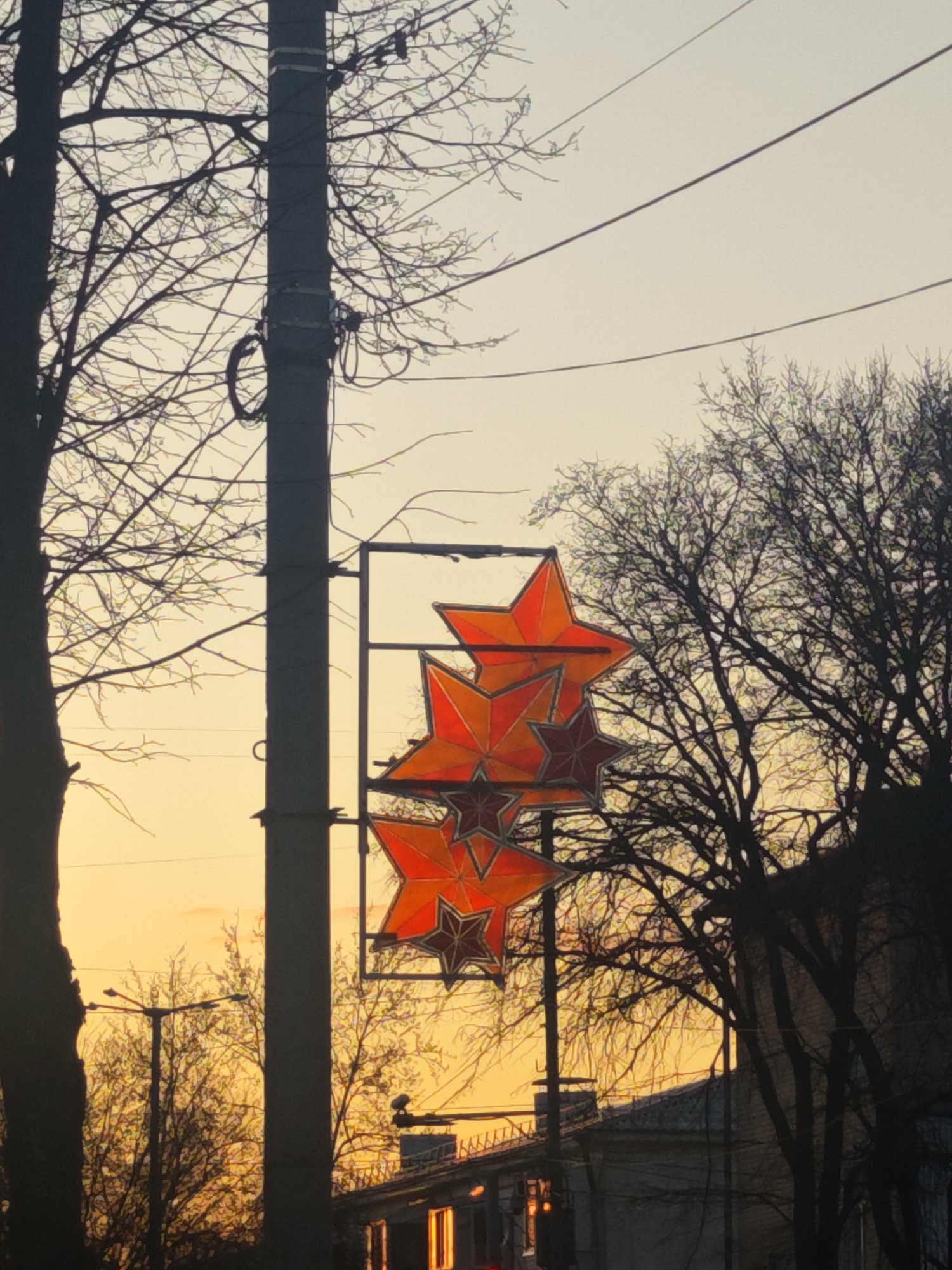 A view in Chaikovsky, Russia features a utility pole between bare trees, with attached, three red star displays hanging on a metal frame. The background consists of a sunset-colored sky and building silhouettes. There are no people present.