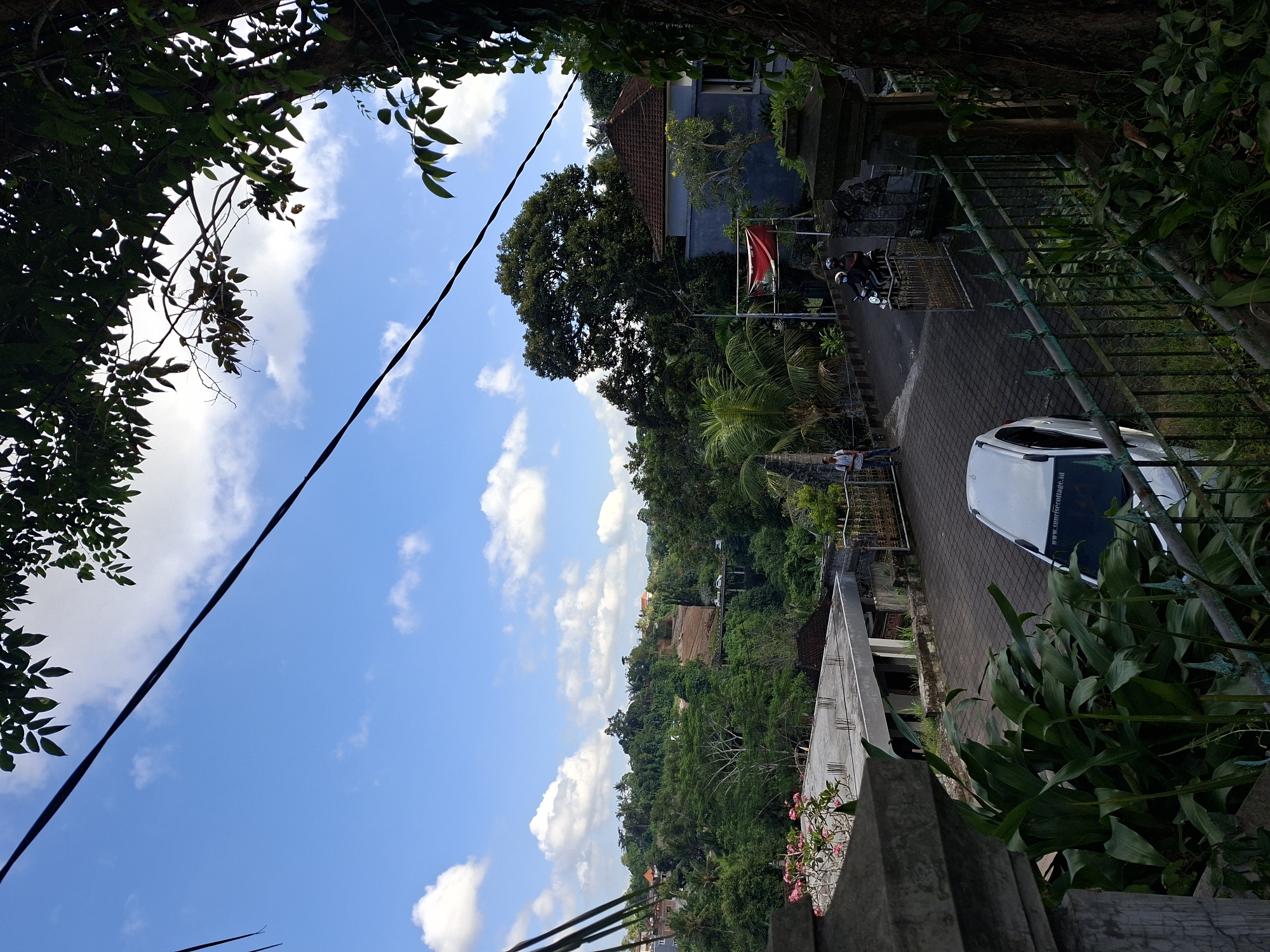 The image depicts a street scene in Banjarangkan, Indonesia, characterized by lush vegetation and a bright, cloudy sky. In the foreground, a gray stone pillar and a dark green metal fence frame the view. Partially visible is the rear of a white car parked on a paved road. Further down the road, a person stands near a gate, and two individuals ride a motorcycle. Buildings with tiled roofs are scattered among dense trees and hills in the background, with a bridge visible in the distance. Overhanging tree branches and a dark power line cross the top of the frame.