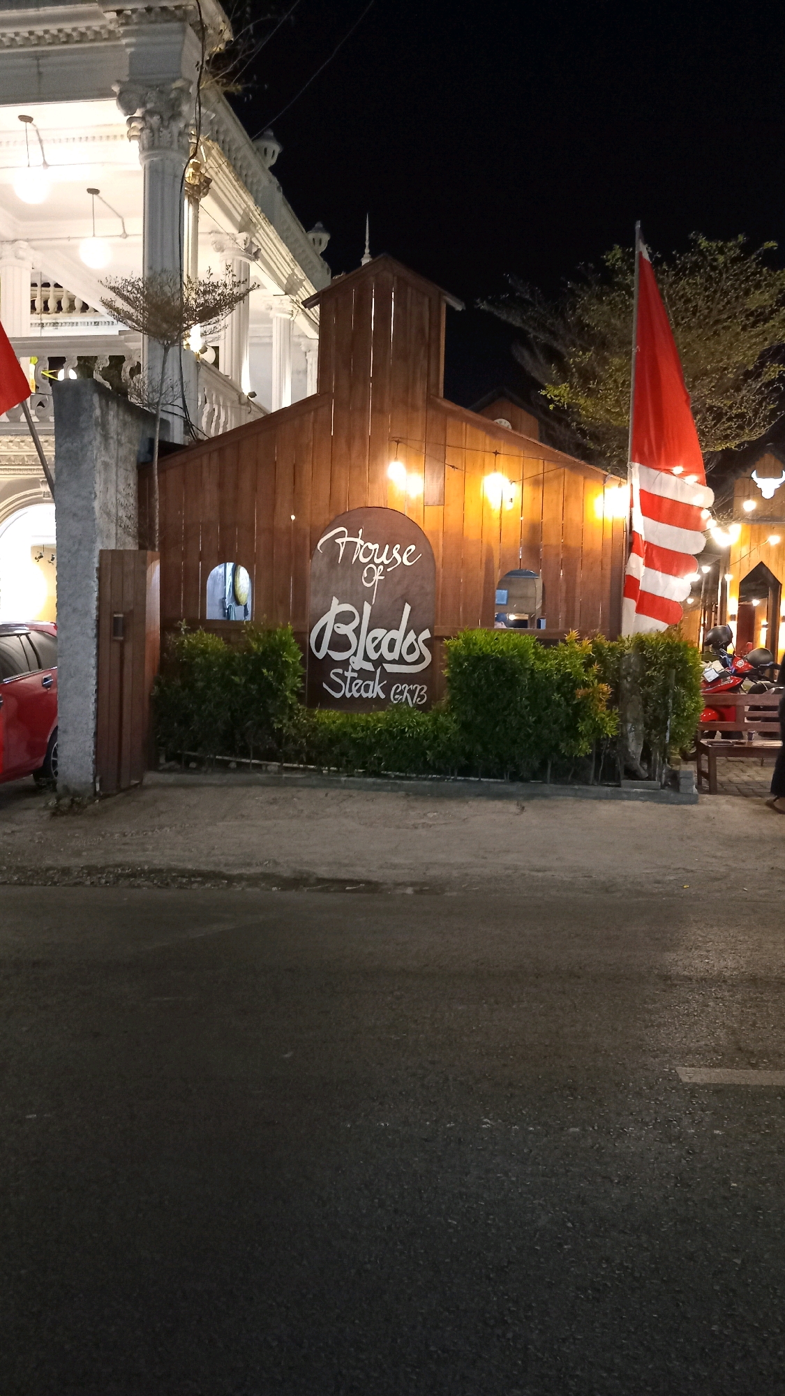 A nighttime exterior shot captures the "House of Bledos Steak" restaurant, situated in Suci, Indonesia. The restaurant features a wooden facade with warm, inviting lights illuminating the establishment. A prominent sign displays the restaurant's name in stylized white lettering. Lush green hedges form a border in front of the building. To the left, a red car is parked next to a gray concrete pillar and a wooden gate. A red and white striped flag is visible on the right side of the frame. The scene is set against a dark night sky.