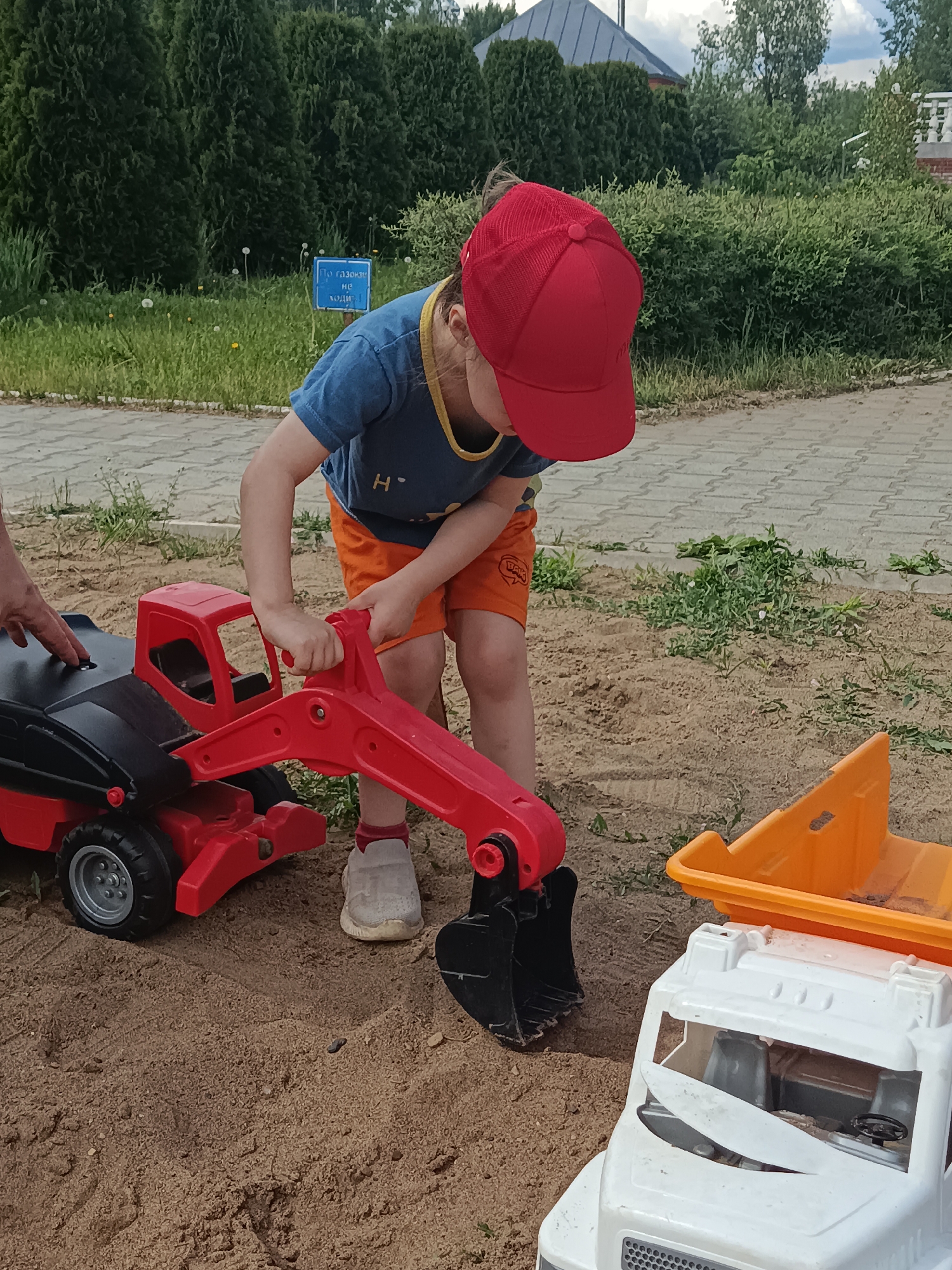 A young child wearing a red baseball cap, blue t-shirt, and orange shorts is playing with toy construction vehicles in a sandbox. The child is operating a red and black toy excavator, while a white and orange toy truck is also present in the sandbox. The sandbox is located outdoors on a paved area, likely a playground or park, as indicated by the paving stones and the presence of trees and bushes in the background. The location is Chaikovsky, Russia. A hand is partially visible on the left side of the image.