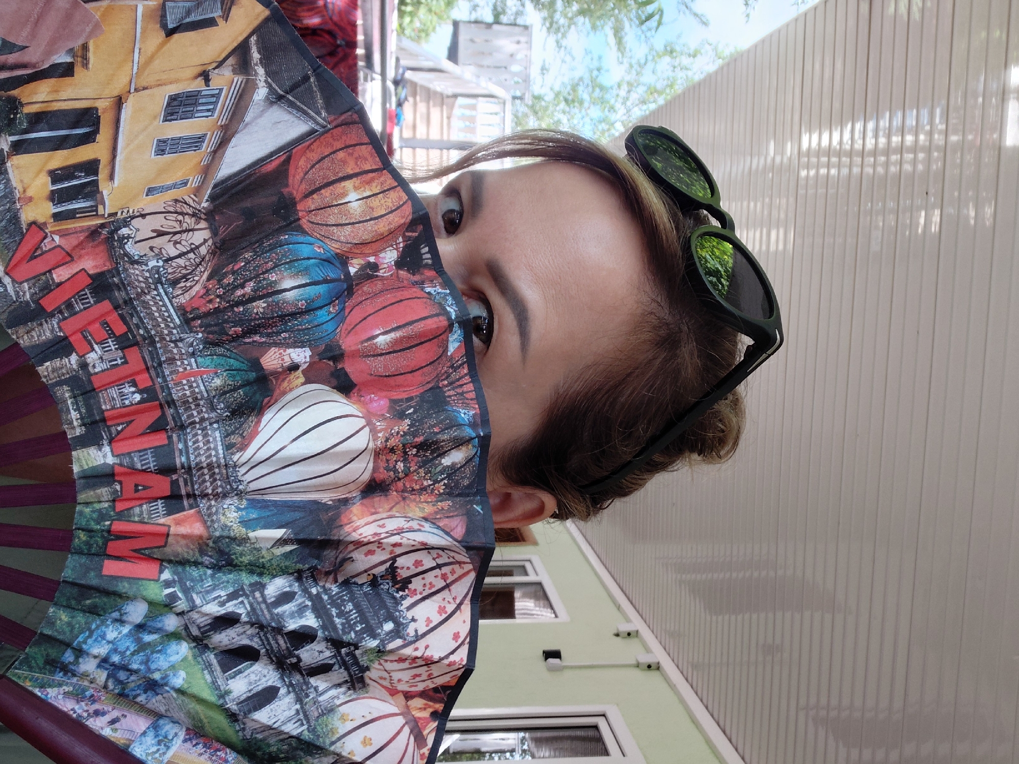 A woman is holding a large, decorative fan in front of her face, with only her eyes and forehead visible. She is wearing dark sunglasses on top of her head. The fan is adorned with images of hot air balloons, traditional Vietnamese architecture, and the word "VIETNAM" in large red letters. The background shows a light-colored, corrugated ceiling, a pale green wall with white trim, and windows. Trees and a wooden structure are visible in the distance. The scene appears to be outdoors, possibly on a porch or patio, in a location that suggests a warm climate. The city of Ақши, Kazakhstan is not depicted in this image.