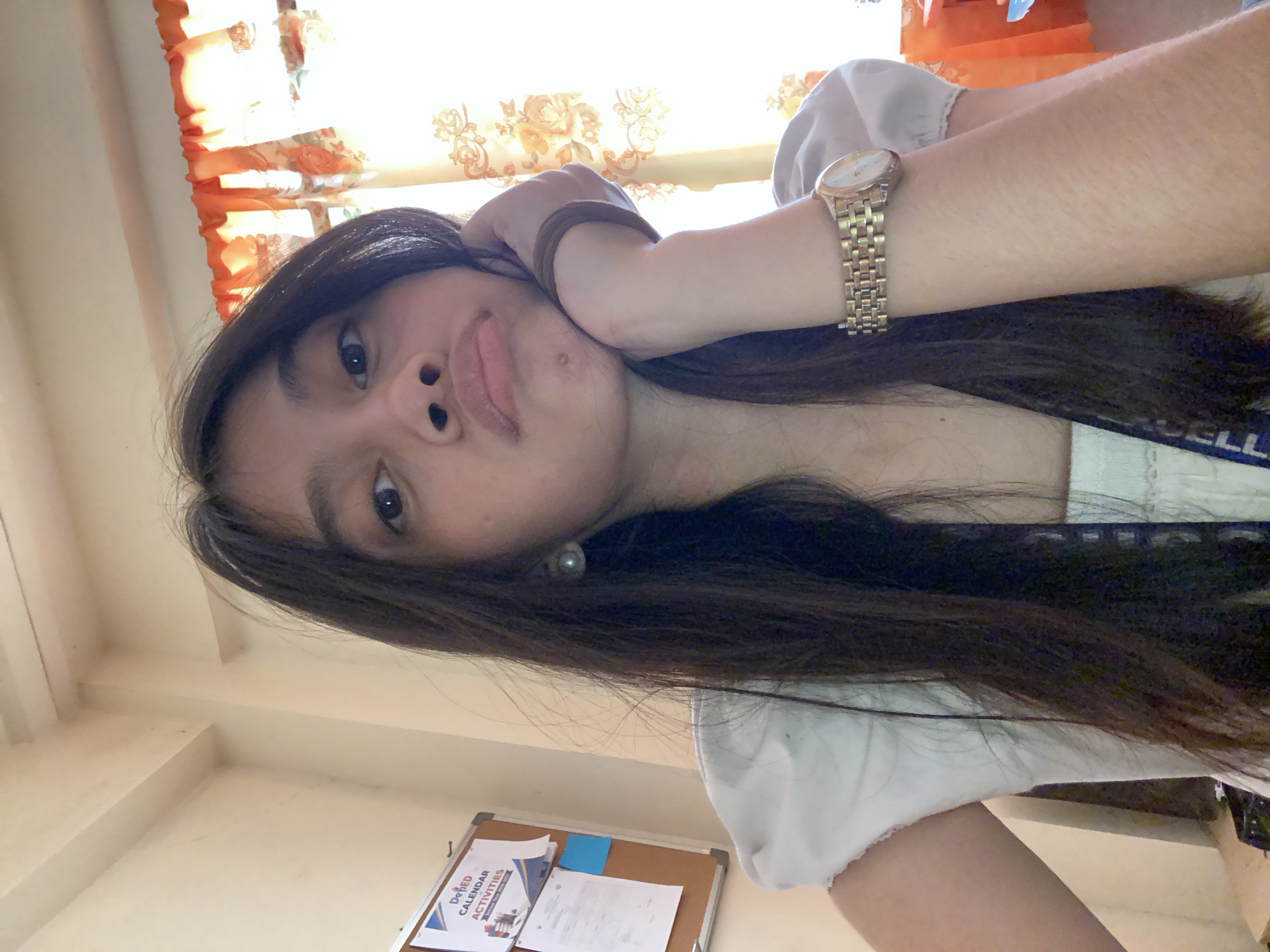 The image is a close-up, selfie-style photograph of a young woman with long, dark hair. She is indoors, and the background suggests a room with a bulletin board on the left wall, displaying papers and a blue sticky note. A window with orange curtains is visible on the right. The woman is resting her chin on her hand and has a slightly pensive or pouty expression. She is wearing a white top with puffed sleeves and a gold wristwatch on her left wrist. The lighting suggests it is daytime. The location is identified as Panabo City, Philippines.