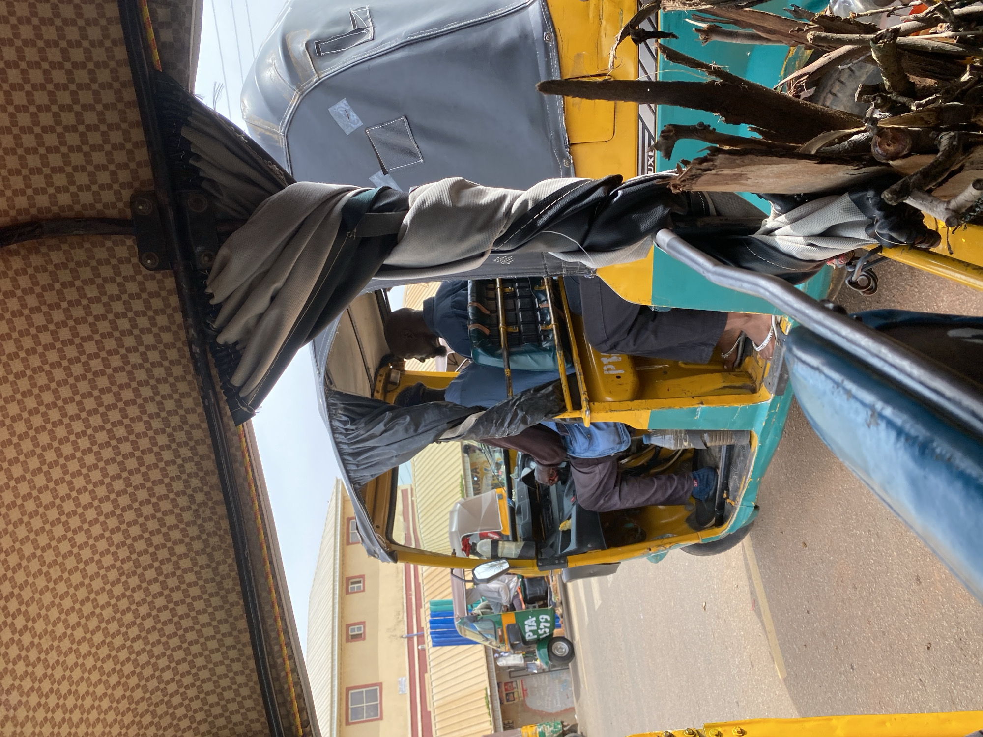 This media file captures a scene from Jos, Nigeria, depicting the interior and immediate exterior of a yellow and blue auto-rickshaw, commonly known as a "Keke Napep" or "Marwa".
The primary subjects are the passengers within the auto-rickshaw. A man, likely the driver, is seated in the front, wearing a dark shirt and dark pants. He appears to be focused on operating the vehicle. Behind him, another person is partially visible, also seated. The auto-rickshaw is loaded with a significant quantity of dried branches and firewood, occupying a substantial portion of the rear compartment. The exterior of the auto-rickshaw is a vibrant yellow and turquoise, typical of commercial vehicles in the region.
The scene is set outdoors, on a paved road, with buildings visible in the background. One building appears to be a light yellow structure with a patterned beige awning, while another is a multi-story building with visible windows and a light-colored roof. The sky is overcast, suggesting a bright but potentially cloudy day.
A notable detail is the patterned fabric lining the interior roof of the auto-rickshaw, which has a checkered design in brown and beige. The metal frame of the vehicle is visible, as are some of the mechanical components. A side mirror is attached to the exterior.
No specific activity or event is explicitly taking place beyond the general context of transportation. The overall impression is one of everyday commerce and travel in a Nigerian urban or semi-urban environment.
Visible text includes "PTA 1579" on a license plate of another auto-rickshaw visible in the background, and partially obscured text on the side of the main auto-rickshaw that includes "UXL".