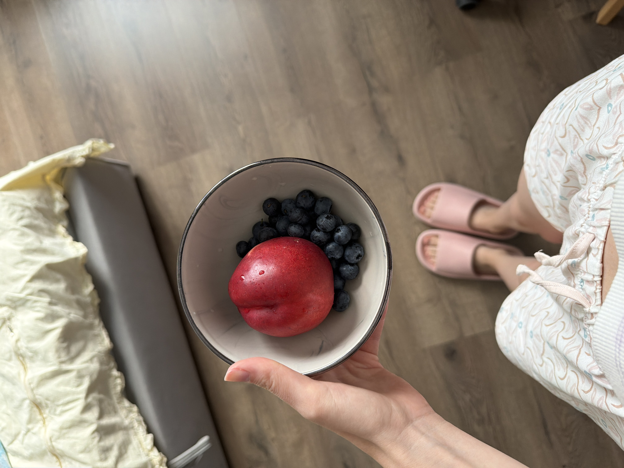 An overhead shot shows a person's hand holding a white and grey bowl filled with blueberries and a single red nectarine. The person is wearing light pink slippers and patterned pajama bottoms. The background features a wooden floor and a bed with a light yellow comforter. The image is a personal, indoor scene, not indicative of any specific city or location.