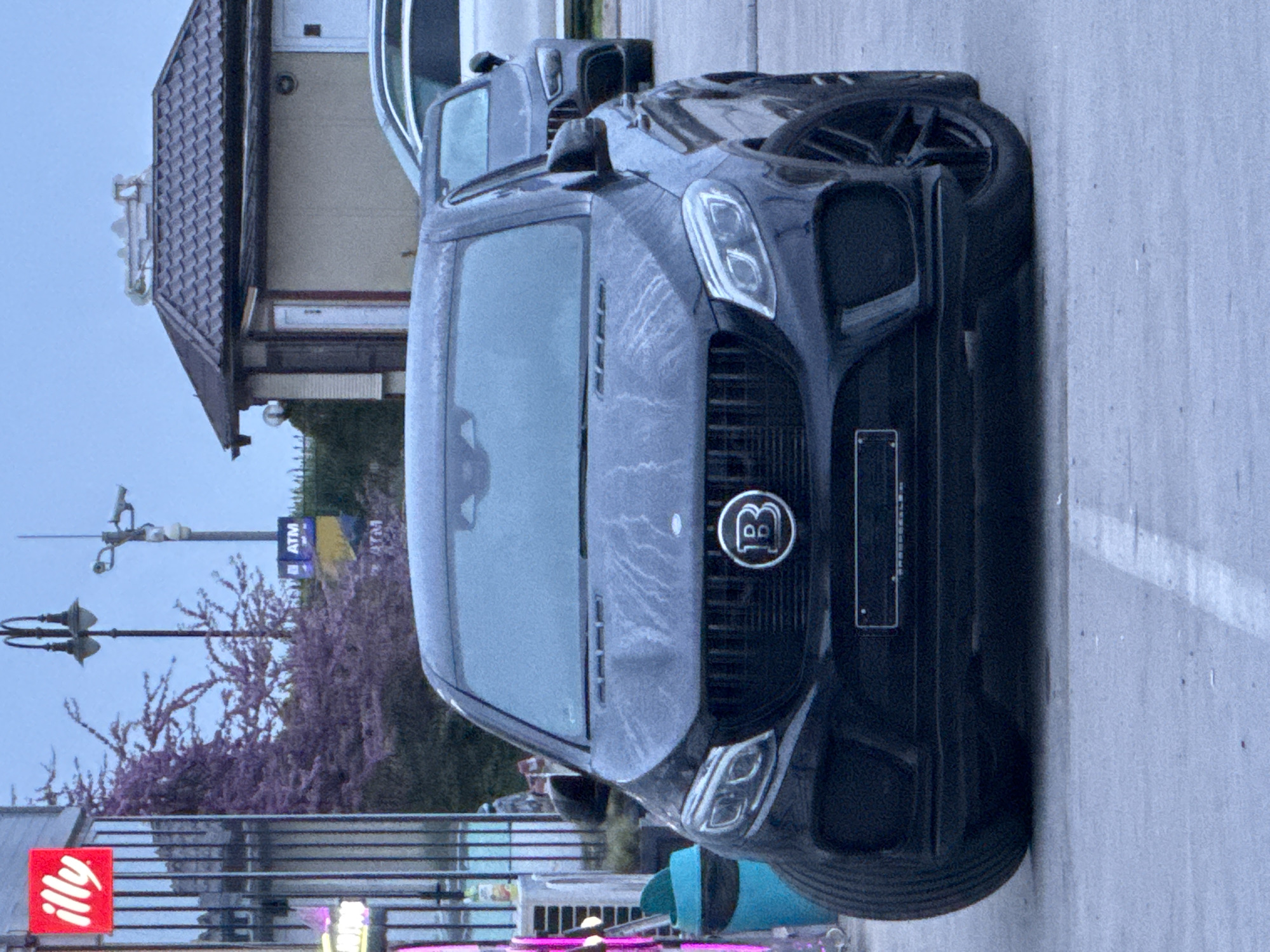 A black Brabus SUV is parked on a concrete surface. The vehicle's front is prominently displayed, showcasing its distinct grill with the "B" emblem. The windshield is covered with a layer of moisture. In the background, there is a building with a gabled roof and light-colored walls. A streetlamp and flowering trees are also visible in the scene. A red "illy" logo is seen on a sign, and an ATM sign can be discerned in the distance. Given the presence of the "illy" logo and based on visual identification, the location is Varna, Bulgaria.