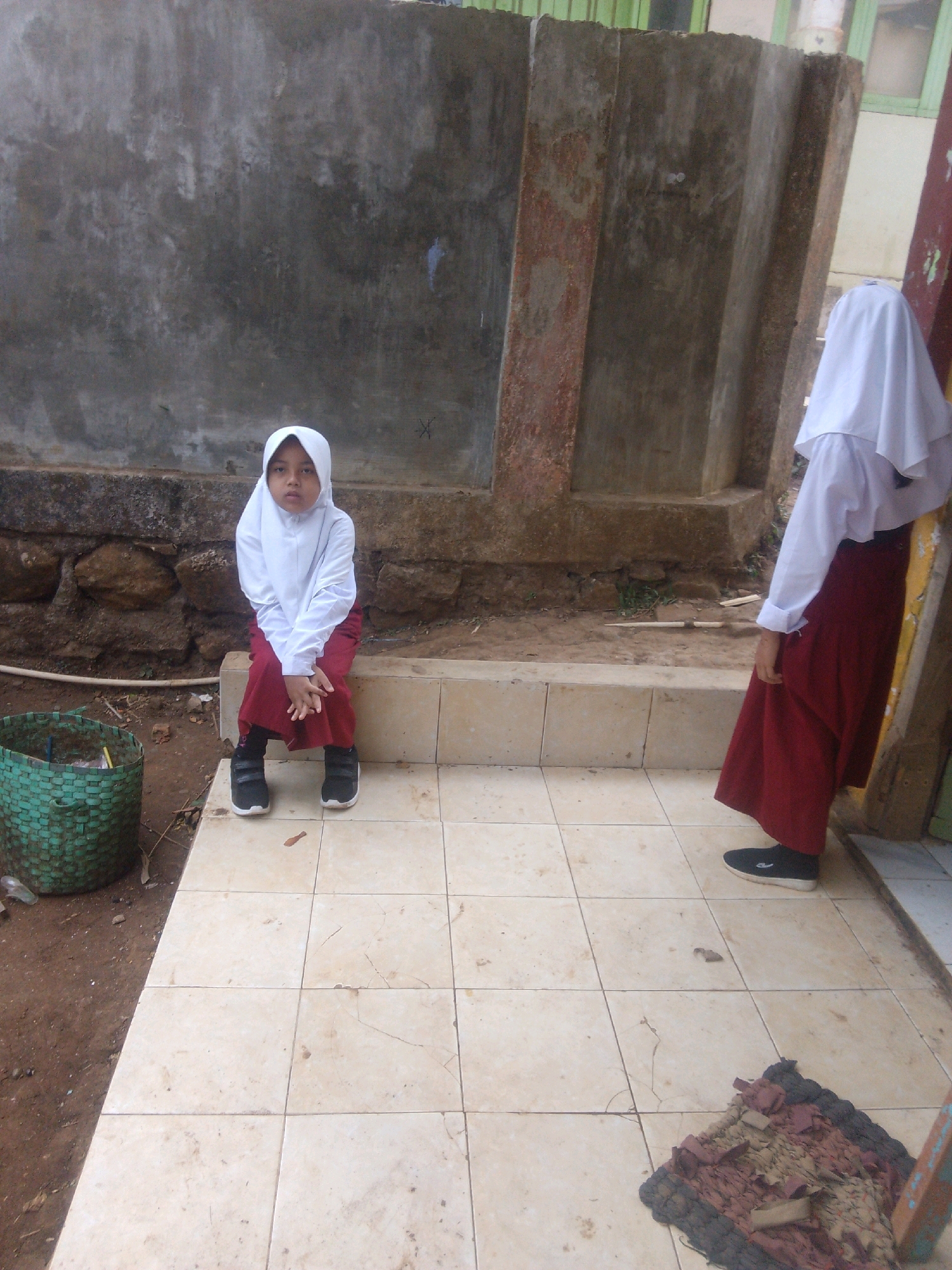 In Betek, Indonesia, a young girl in a white hijab and maroon skirt sits on a concrete ledge. To her right, another individual, also wearing a white hijab and maroon skirt, stands. A green woven basket is positioned to the left of the seated girl. The ground is covered with beige tiles, and in the foreground, a small, colorful rug lies on the tiles. The background features a weathered concrete wall and some vegetation.