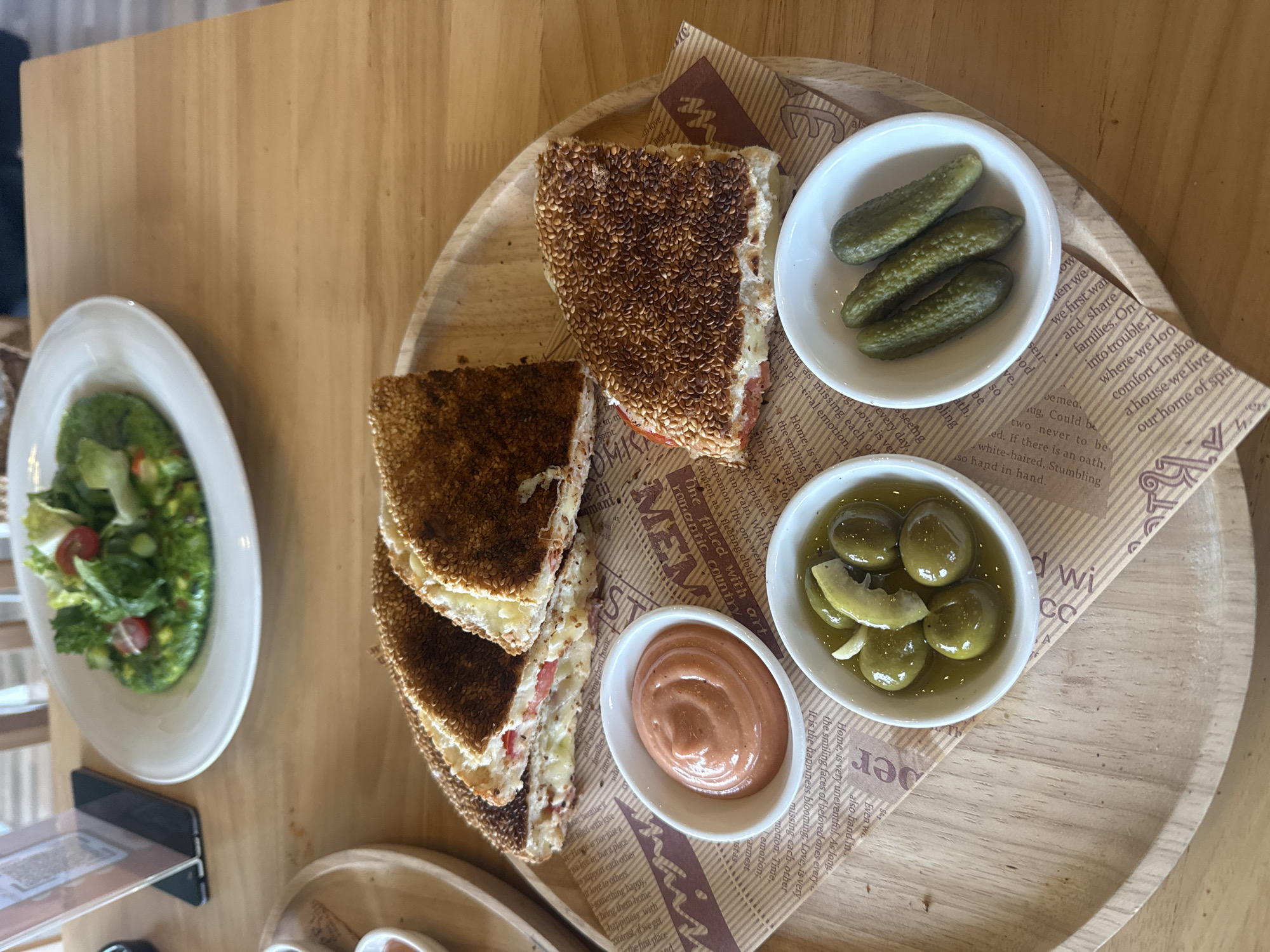 A top-down shot shows a wooden table with two plates of food and some cutlery. In the foreground, a circular wooden tray holds a toasted sandwich cut into triangles, with a side of olives, pickles, and a pinkish sauce. The sandwich is coated in sesame seeds and appears to be filled with cheese and tomato. The olives are whole and pitted, while the pickles are small gherkins. The sauce is in a small white bowl and has a swirl pattern. Behind the tray, a white plate holds a salad with lettuce, cherry tomatoes, and what appears to be a green spread underneath. A black tablet stands upright on the table to the left. The environment suggests a casual dining setting in a restaurant.