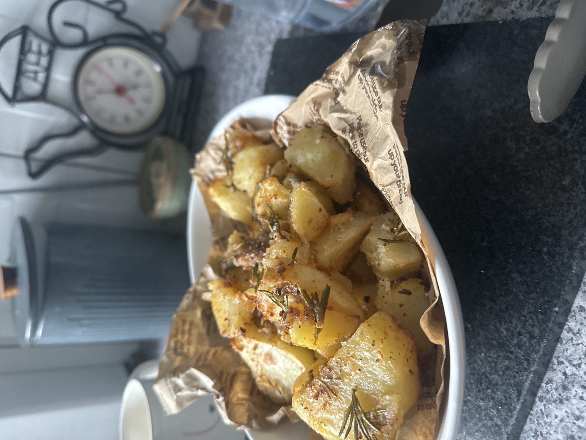 A close-up, eye-level, indoor shot shows a white bowl overflowing with golden-brown roasted potato wedges, seasoned with herbs such as rosemary and coarse salt. The potatoes are served on crinkled brown paper, possibly parchment, which is partially tucked into the bowl. In the background, a light grey, ridged cylindrical canister with a wooden lid stands to the left. Further to the right, a decorative black metal teapot-shaped clock with Roman numerals is visible, and to its left, a small green bowl. The surface beneath the bowl appears to be a dark, speckled countertop. The lighting suggests an ambient indoor environment. The location is not specified in Macclesfield, United Kingdom.