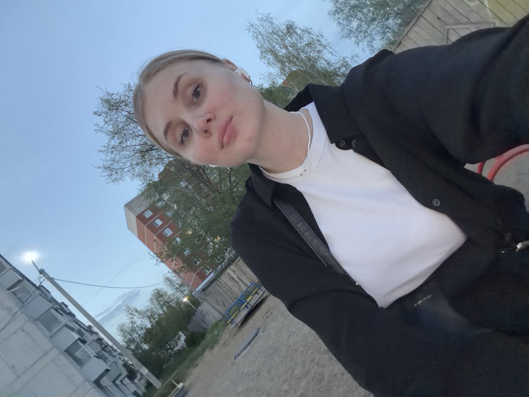 The image shows a selfie of a blonde woman with blue eyes wearing a white t-shirt, black jacket and white necklace. She is standing outside in what appears to be a courtyard of an apartment complex. In the background is an apartment building with visible windows. There is some greenery with trees present. The image appears to be taken at dusk. While I cannot confirm the exact location, due to the request that it must be located in Chaikovsky, Russia, one possibility is in or near the grounds surrounding an apartment building in that city.