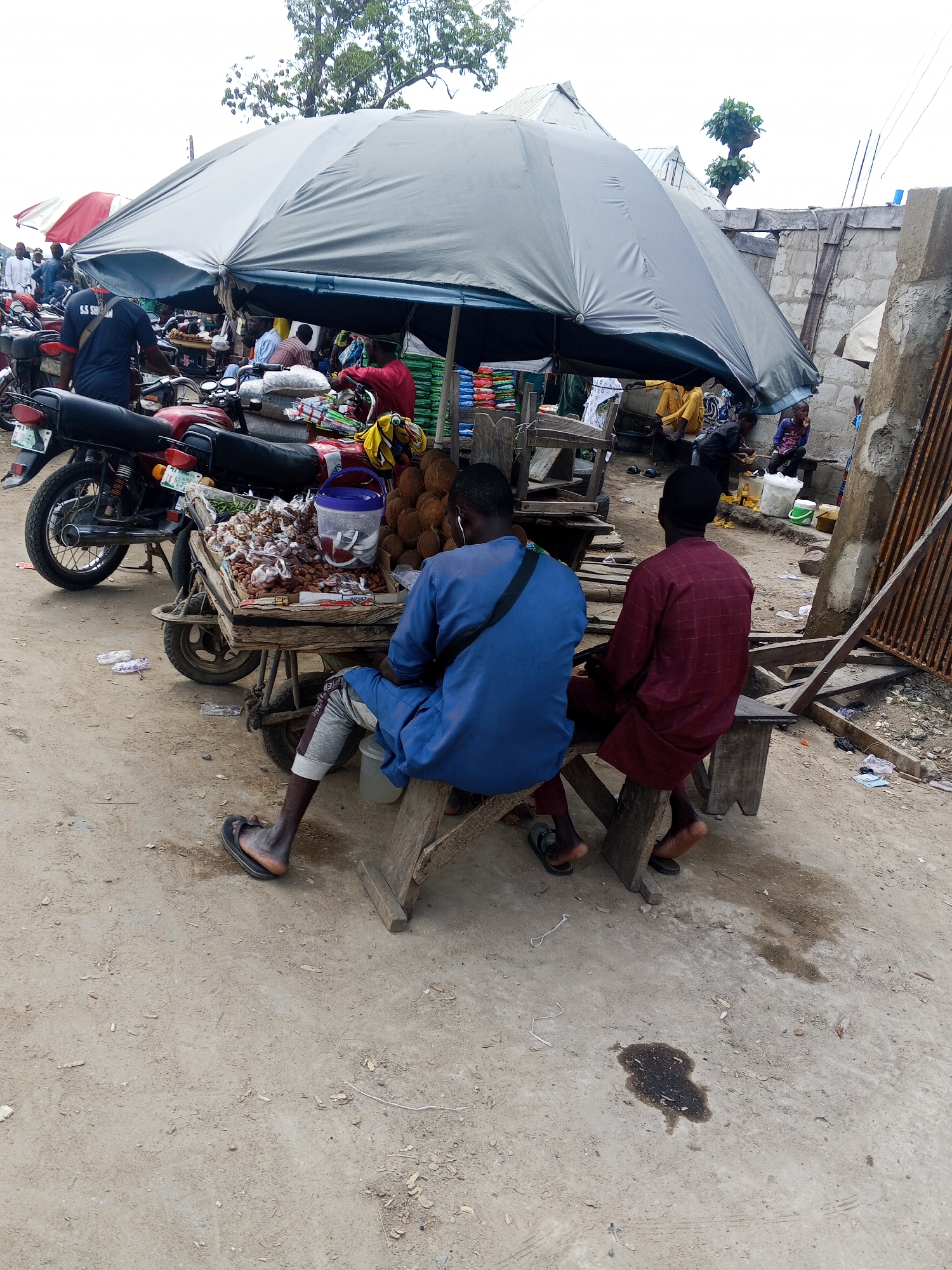 A daytime, eye-level, outdoor shot captures a bustling marketplace scene in Zango-ndaji, Nigeria, dominated by a large, grey tarp-covered stall. Two men are seated on makeshift wooden benches in the foreground, facing away from the camera, under the shade of the stall. The man on the left wears a blue tunic and grey trousers with flip-flops. The man on the right wears a maroon patterned tunic and black sandals. In front of them, a wooden cart is laden with produce, including what appears to be dried beans or nuts, some bagged, and a blue plastic container. A stack of coconuts is visible to the right of the cart. In the background, several motorcycles are parked, and other market-goers are visible, some blurred or in shadow, suggesting ongoing commerce. The ground is a mixture of dirt and sand, with scattered debris and a dark stain suggesting a liquid spill. Structures, possibly made of concrete blocks and corrugated metal, form the backdrop of the market.