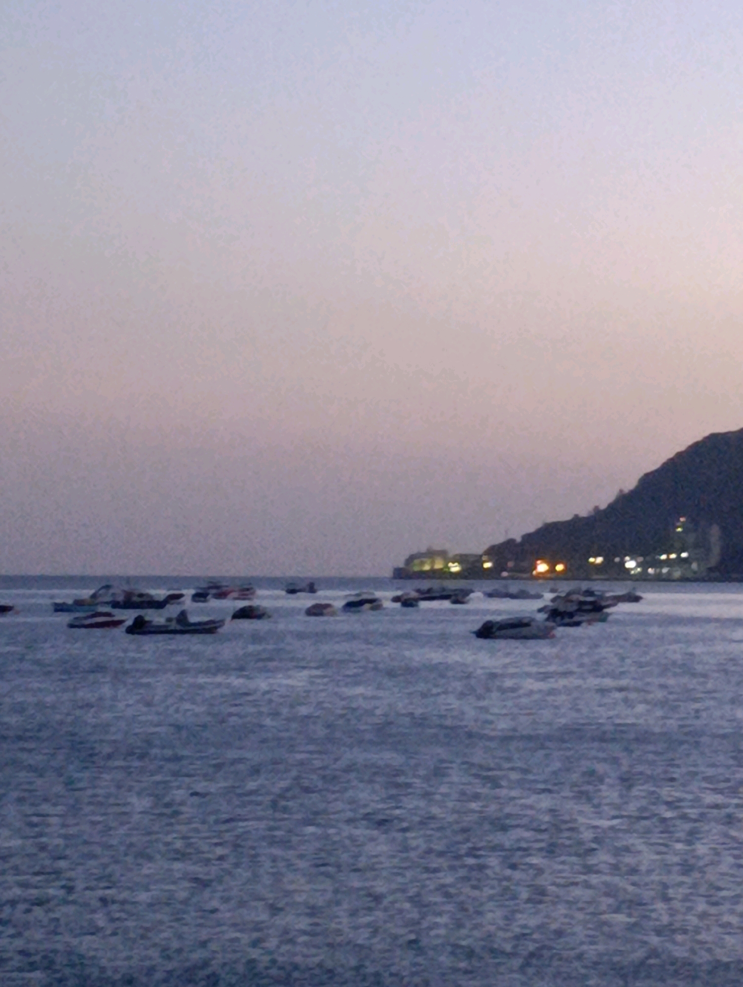 An image depicts a body of water with numerous small boats anchored. In the background, a dark, sloped landmass rises, featuring lights from buildings along its edge. The sky above is a gradient of pale purple and pink, suggesting either dawn or dusk. The water is choppy with visible ripples. The scene appears to be a harbor or coastal area in Setúbal, Portugal, given the characteristic topography and lighting. No people are discernible in the image.