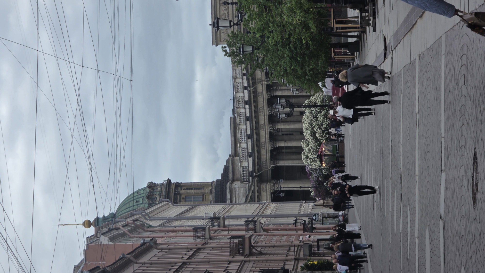 The image depicts a street scene in Saint Petersburg, Russia. A paved walkway is lined with buildings on the left and a classical building with columns on the right. People are walking along the walkway, some in groups. The building on the left has a green dome and a gold spire. Overhead, a network of wires is visible against a cloudy sky. The architecture is ornate, with detailed facades and columns. Along the street, there appear to be trees, light posts, and possibly floral decorations.
