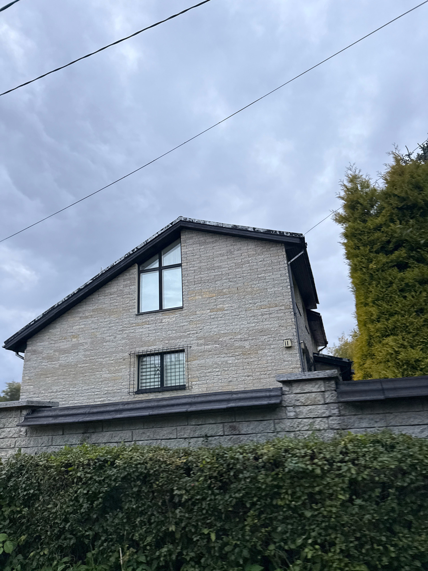 The image shows a stone-clad house with a dark-pitched roof. A large, angular window is visible on the upper floor of the house, and a smaller, rectangular window with a metal grate is on the lower floor. The house is bordered by a stone wall, which is topped by a dark gutter or capping. In front of the wall is a dense green hedge. A tall, evergreen tree is situated to the right of the house. The sky is overcast and cloudy. The scene appears to be in a residential area, possibly in Tallinn, Estonia, given the architectural style and the presence of utility lines overhead. No people are visible in the image.